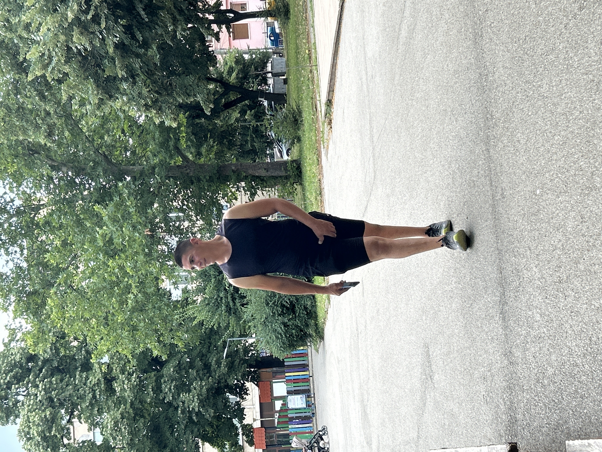 A young adult male is walking on an asphalt path in an urban park setting in Vratsa, Bulgaria. He is wearing a black sleeveless athletic shirt and black athletic shorts, with athletic shoes on his feet. He is holding a mobile phone in his right hand, with his left hand in the pocket of his shorts. In the background, there are trees, buildings, a playground with a colorful fence, and parked cars. The sky is overcast.