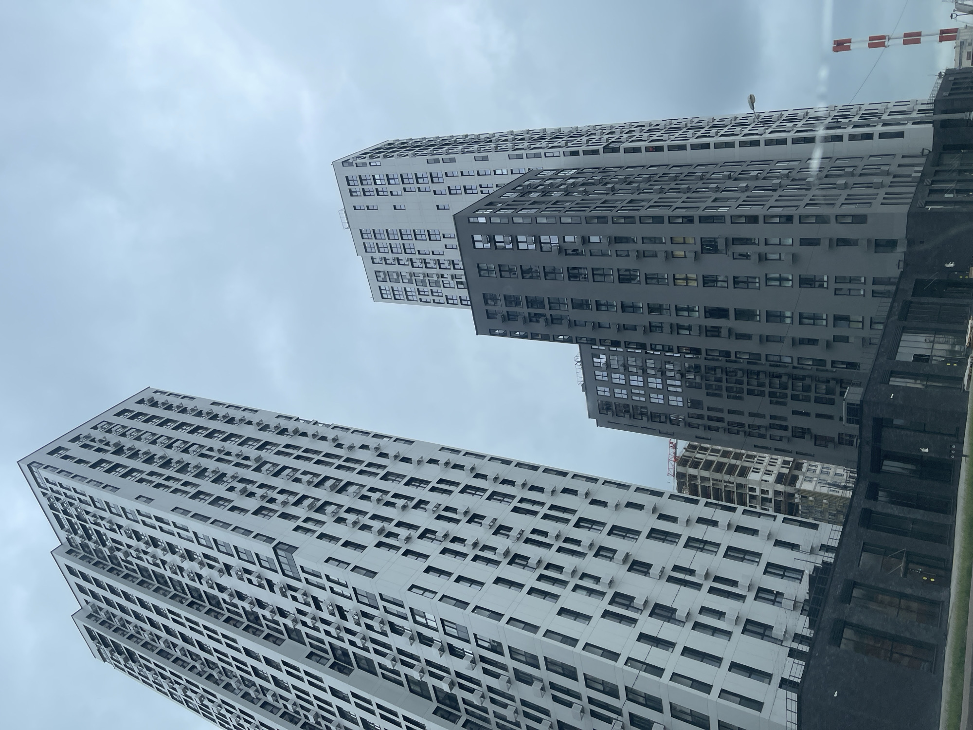The image depicts a cluster of modern high-rise residential buildings under a cloudy sky, likely in Moscow, Russia. The tallest building, on the left, is a white skyscraper with a repetitive facade of dark-framed windows and numerous protruding air conditioning units. To its right, a slightly shorter, dark gray skyscraper shares a similar design with rows of windows and air conditioning units. In the background, another white skyscraper, taller than the gray one but shorter than the leftmost white building, is visible, also featuring a grid of windows. Further back, a building under construction with exposed concrete and scaffolding can be seen. The foreground shows the dark, brick facade of a lower building with large, dark windows. A lamppost stands to the right of the dark gray skyscraper, and a red and white striped industrial chimney is visible in the far right background. No people are discernable in the image. The overall impression is one of dense urban development with a focus on residential architecture.