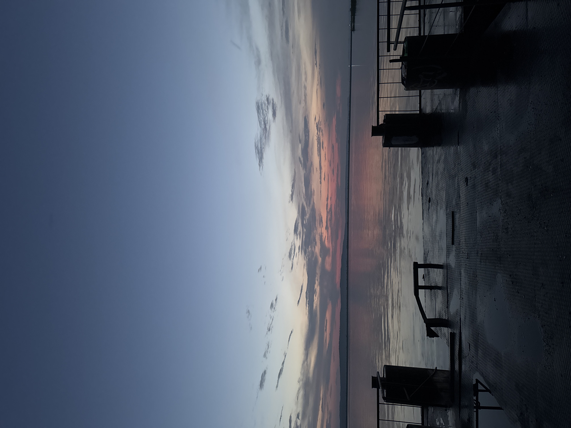 The image depicts a serene sunset over the Kama River in Chaikovsky, Russia. In the foreground, a wet, metallic pier stretches into the water, reflecting the soft hues of the sky. Two large cylindrical bollards stand on the pier, serving as mooring points. A metal railing runs along the right side of the pier, and a set of metal steps leads down to the water. The water itself is calm, with gentle ripples reflecting the colors of the sunset. In the distance, a tree-lined shore is visible, fading into the hazy horizon. The sky is a gradient of blues and pinks, with scattered clouds adding texture and depth to the scene. The overall atmosphere is peaceful and tranquil, evoking a sense of quiet contemplation.