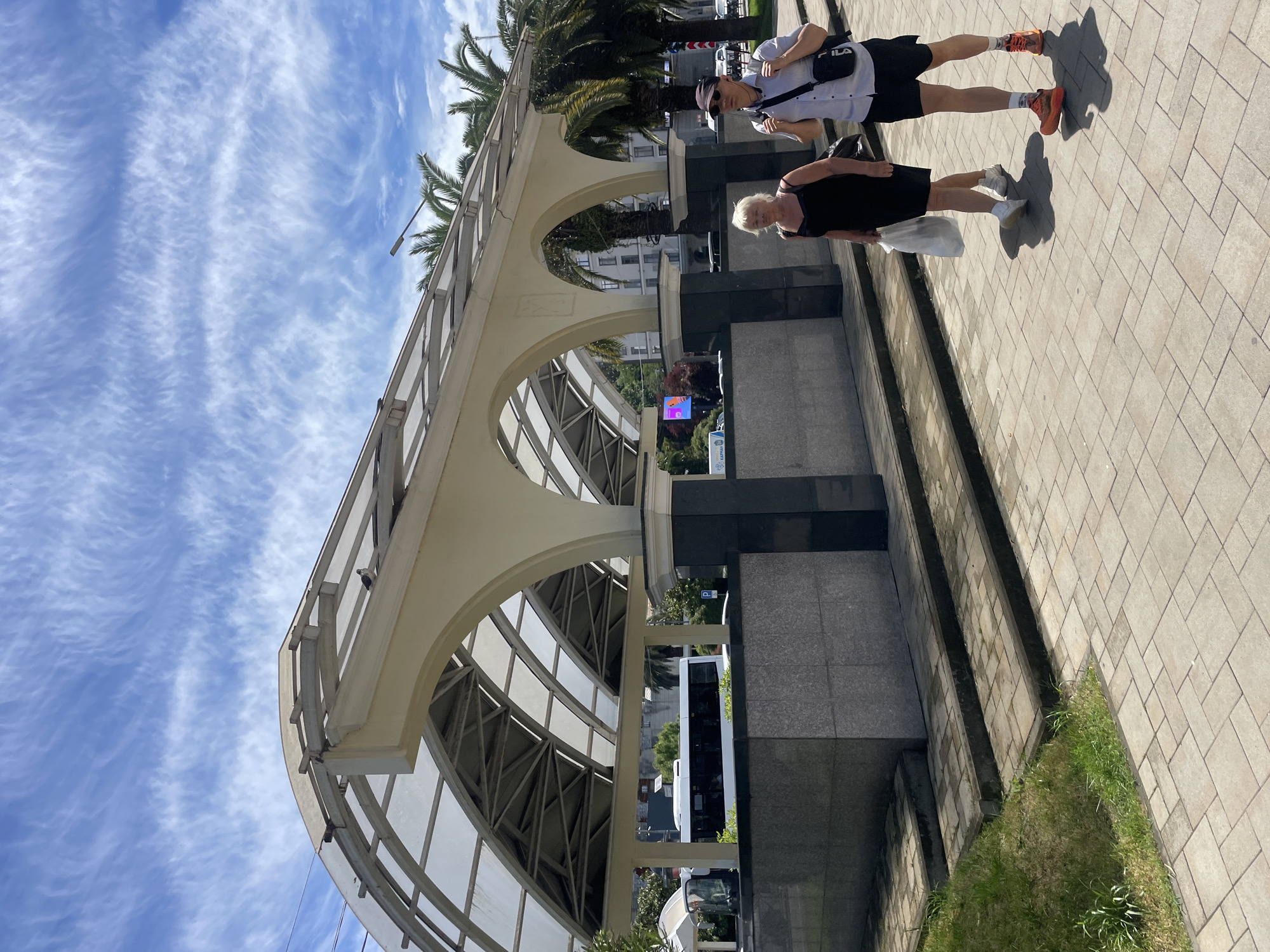 The image captures a scene in Sochi, Russia, featuring a pedestrian walkway with arched supports. Two people are walking on the paved sidewalk. A young man wearing a white shirt, black shorts, a hat, and orange shoes has a black shoulder bag. An older woman in a black dress is also walking. In the background, palm trees are visible and a building can be seen through the arches. A white bus is parked beneath the walkway. The sky is blue with cirrus clouds.