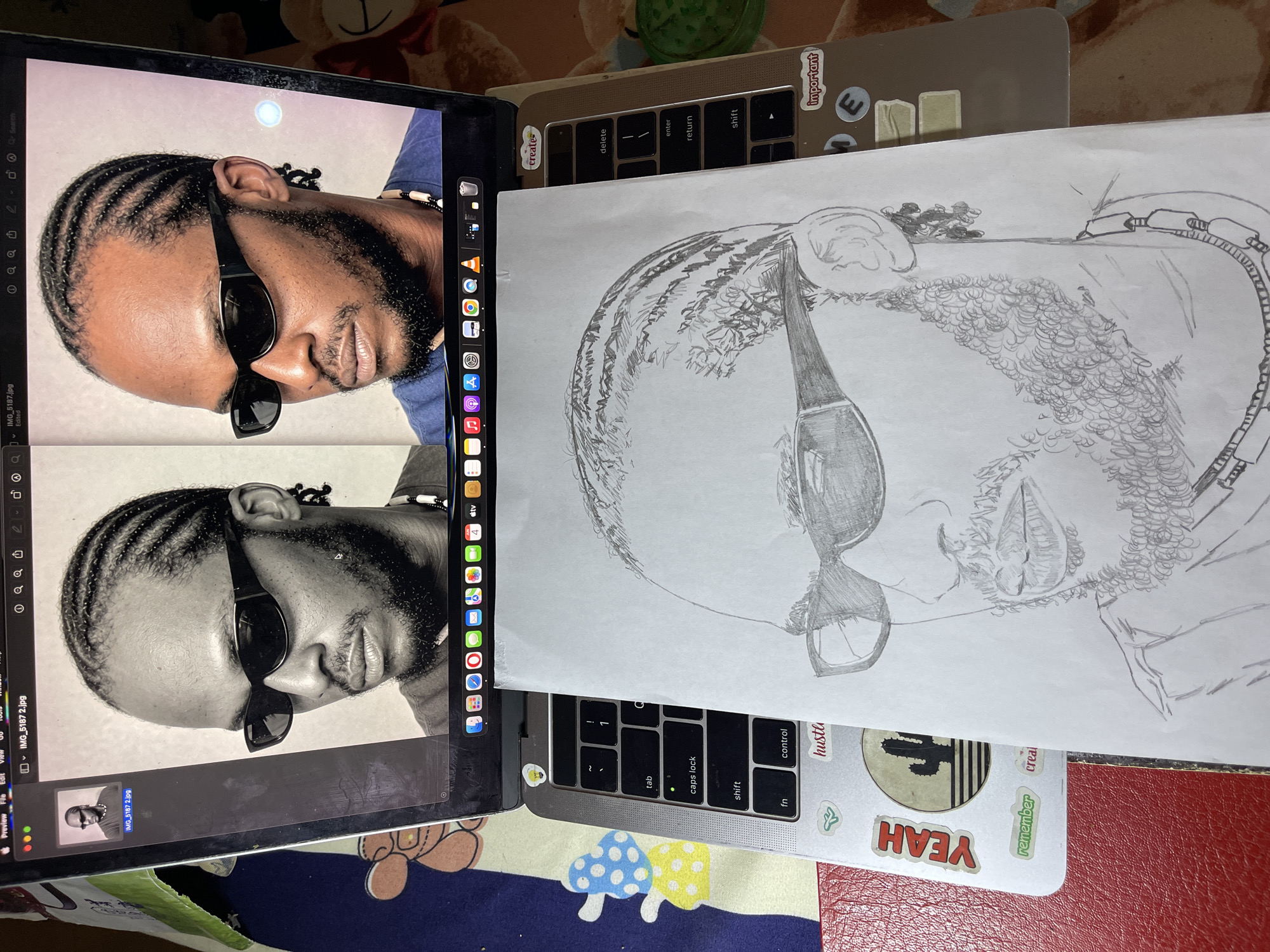 To accurately describe the media file, let's break down the visible content and context: **Subjects:** The primary subjects are a person (male) and two forms of media portraying him: digital images displayed on a laptop screen and a hand-drawn pencil sketch on paper. **Objects:** A closed silver Apple MacBook Pro laptop (partially open on the right, providing a surface for the sketch). The main display of an open laptop (possibly another MacBook or an iPad) is visible on the left, showing two images of the man. A sheet of white paper holds the pencil sketch. Various items are on the laptop's keyboard area, including several stickers: "important," "create," "hustle," "remember," "creal," and "YEAH," along with a round black and white sticker featuring a cactus. A green bottle or container is in the upper background, and a brown plush toy (possibly a bear) is visible behind the laptop. The surface beneath the laptop appears to be a patterned textile or mat. **Activity or Event Taking Place:** The arrangement suggests an artistic process or display. The two digital images of the man on the laptop screen are likely reference photos for the pencil sketch. One image is in color, and the other is a black-and-white version of what appears to be the same person. The pencil sketch closely resembles the man in the digital images, indicating an artist's rendition. It's possible someone is either in the process of drawing, or showcasing a completed drawing alongside its inspiration. **Notable Details:** The man in the images and sketch is depicted with dark skin, braided hair (cornrows), a beard, and sunglasses. He is also wearing a beaded necklace. The digital images show a direct, slightly angled gaze. The lighting appears to be ambient indoor light, possibly from a window or overhead fixture, with a bright circular reflection on the upper digital image. The sketch features detailed shading on the face, hair, beard, and sunglasses. The overall scene is casual and personal, likely a workspace. **Visible Text:** * On the left laptop screen (menu bar/sidebar, partially visible): "Preview", "File", "Edit", "View", "Tools", "Window", "Help". Also filenames "IMG_5187.jpg" and "IMG_5187 2.jpg", "Edited", "tv". Several application icons are visible on the dock, including what appear to be Adobe Illustrator and Google Chrome. * On the physical laptop's keyboard/palm rest area, various stickers are visible: "important", "create", "hustle", "remember", "creal", and "YEAH". Standard keyboard keys like "tab", "caps lock",