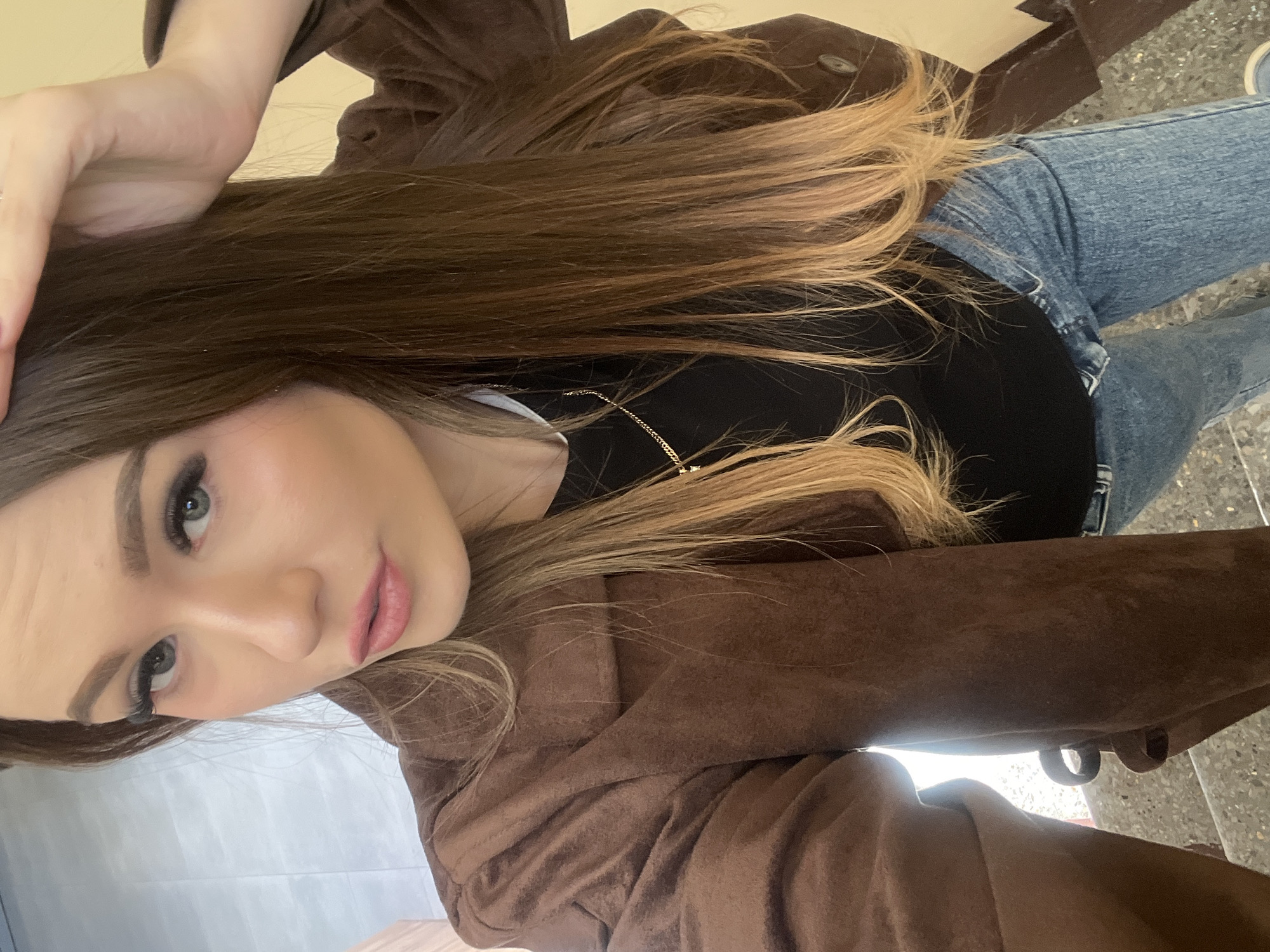 A young woman takes a selfie while wearing a brown suede jacket, a black t-shirt, and blue jeans. She has long brown hair with blonde highlights and is wearing makeup. The background appears to be an indoor setting with a tiled floor. The location is unknown and cannot be determined.