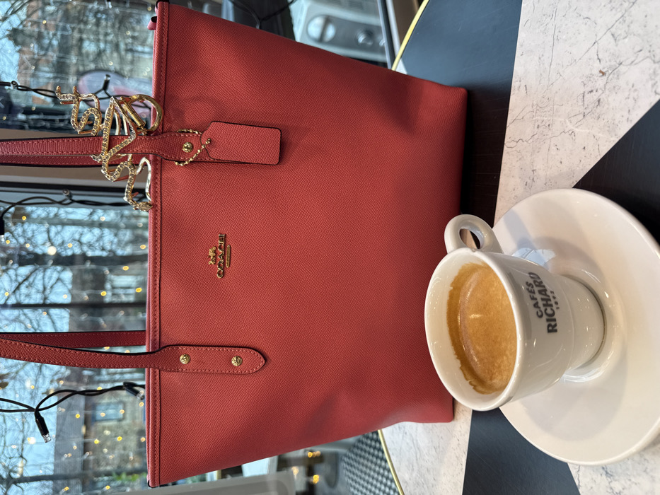 The scene is set indoors, likely within a café in Arcueil, France, looking out through a large window. In the foreground, a coral-red (or salmon-pink) textured leather Coach tote bag stands upright, centered on a table. The bag features gold-toned hardware, including its brand logo centrally placed, two small studs on the front strap attachment, and a small hanging tag of the same material. A gold-toned, serpentine-shaped accessory adorned with small clear stones is clipped to the bag's right strap. To the left and slightly in front of the bag, a white ceramic espresso cup with "CAFÉS RICHARD 1892" printed in black, sits on a matching saucer. The cup contains dark brown liquid with a light brown crema layer. The table surface displays a distinctive pattern of alternating black and white marble-patterned triangles, bordered by a gold-colored edge. The background, visible through the window, is out of focus, showing bare tree branches interspersed with numerous small, warm-white string lights. Faint outlines of distant structures are discernible. To the far left, a small portion of a black and white checkerboard tiled floor is visible. To the far right in the background, a blurred white appliance, possibly an air conditioning unit, is present. No individuals are visible.