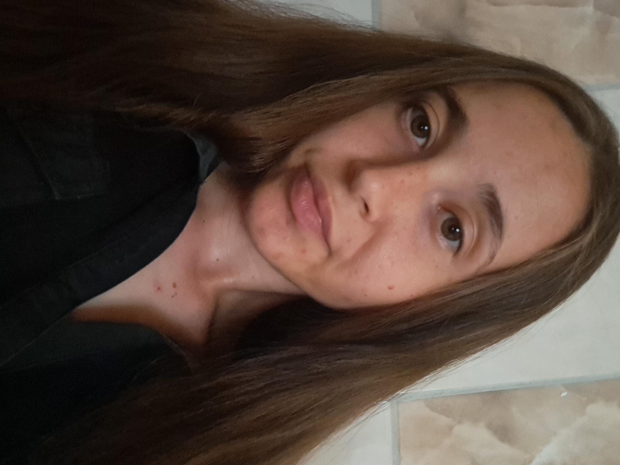 A close-up, front-facing shot shows a young person with long, brown hair and light skin. The person has brown eyes, light eyebrows, and a few freckles scattered across their face. Their lips are slightly parted, and they are wearing a dark-colored collared shirt. The background appears to be a tiled wall with beige and white tones. There is no indication of the location being in Vypasne, Ukraine.