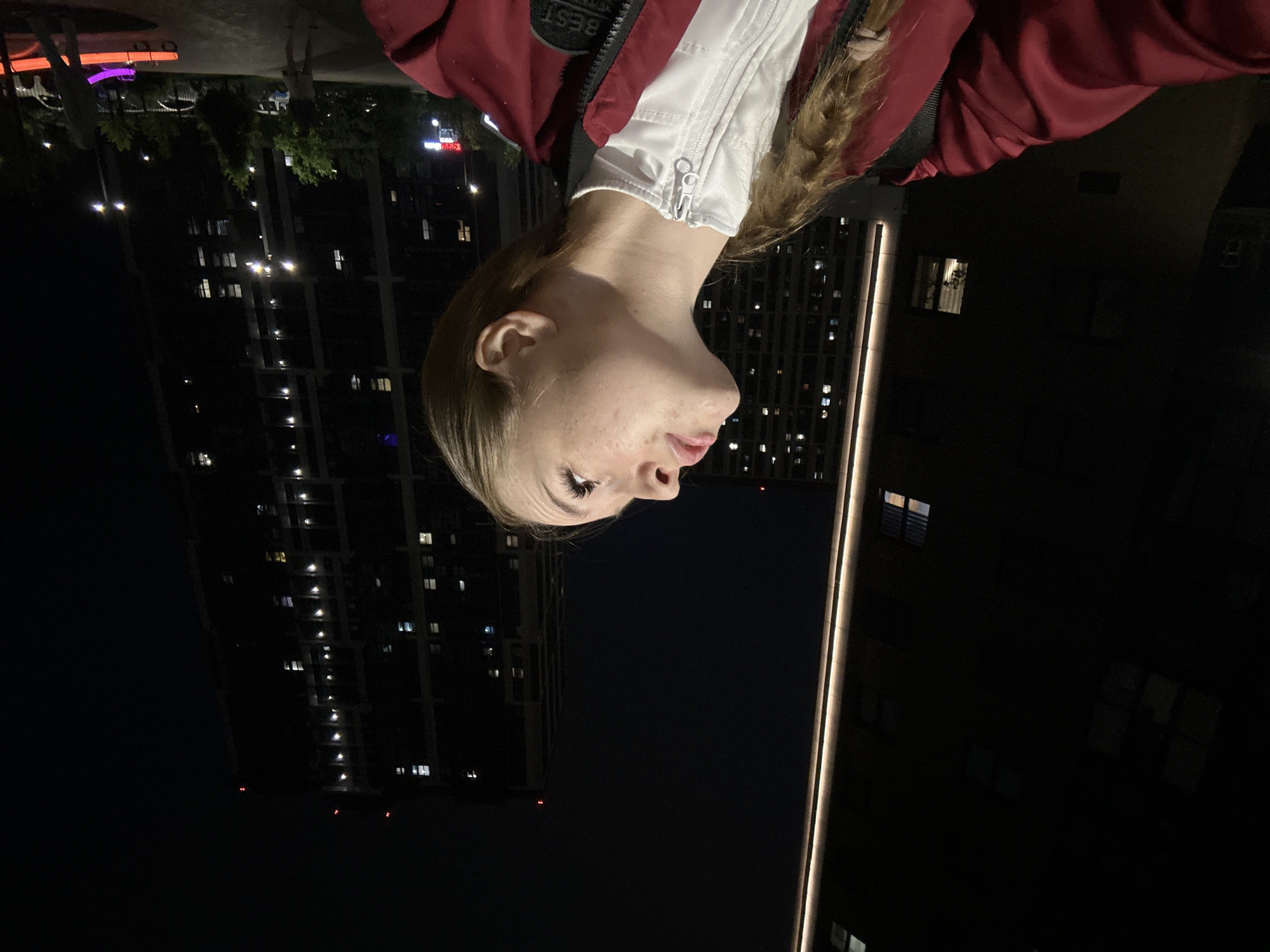 A tall, modern building with many lit windows stands in the darkness of the Unknown, Unknown city at night. A blonde woman in a red jacket is facing the viewer.