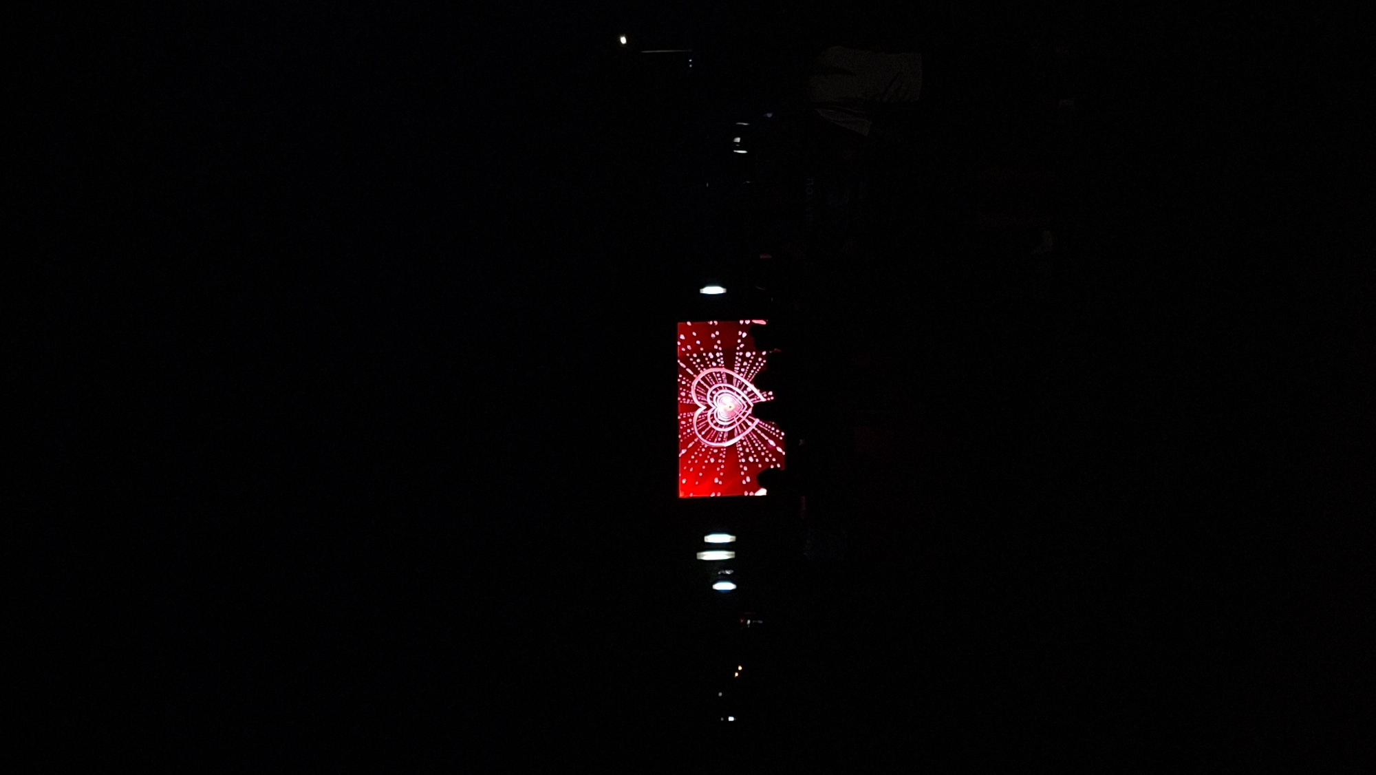 The image captures a night scene where a large LED screen displaying a stylized red heart with radiating white dots and concentric circles is visible. A crowd of silhouetted figures stands in front of the screen, appearing to be watching it. The environment is dark, suggesting it's outdoors in a public space within the city of Myadzyel, Belarus. Streetlights or other ambient lighting are faintly visible in the background, creating a contrast with the illuminated screen. The overall atmosphere is that of an event or gathering taking place at night.