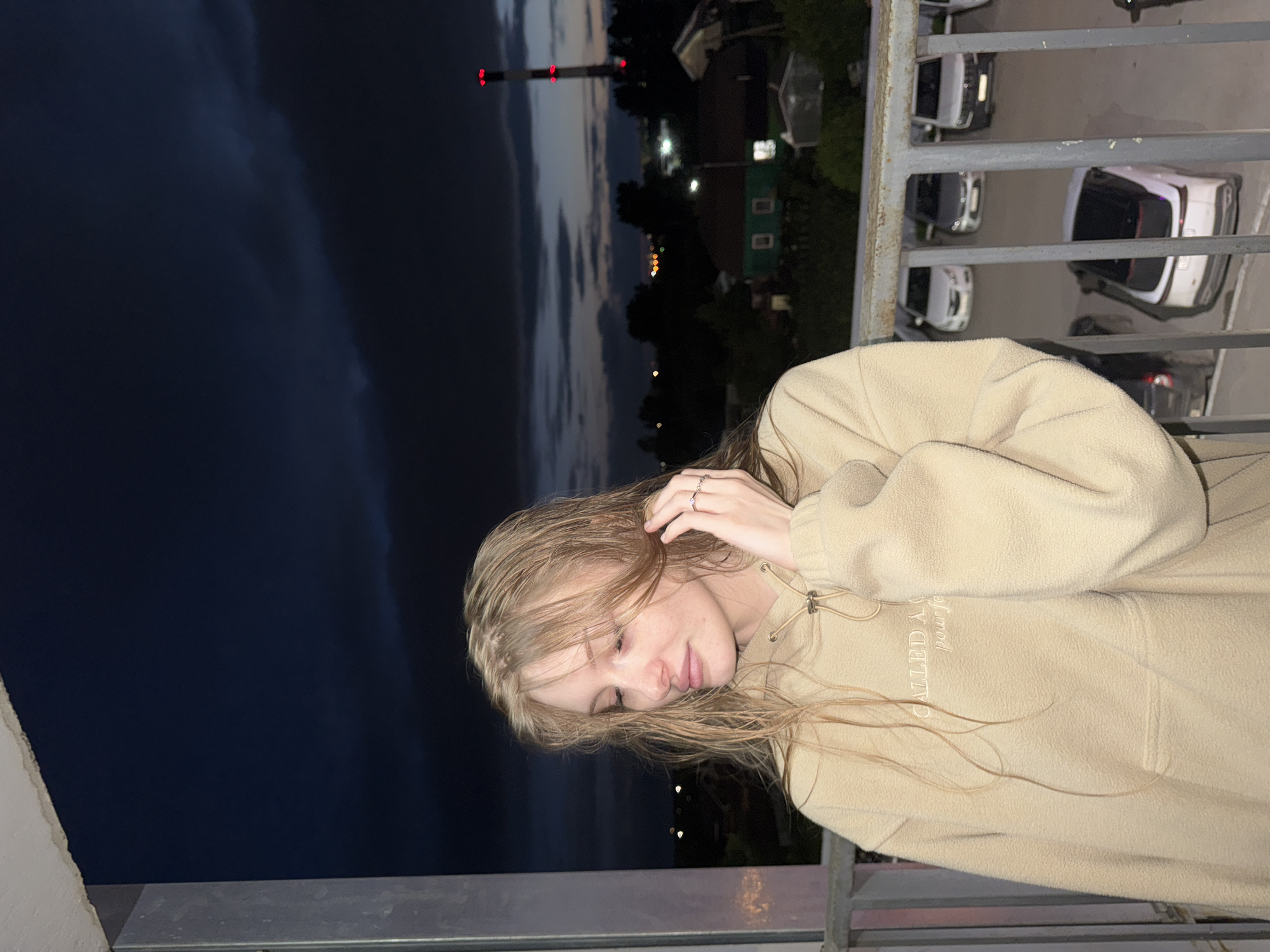A young woman with blonde, wet hair and fair skin is standing on a balcony at dusk. She is wearing a tan, fleece hoodie with embroidered text on the chest. Her right hand is touching her hair, and she is wearing rings on her fingers. She is looking down and to her left. Behind her, the sky is dark blue with some clouds. In the distance, there are houses with lit windows, trees, and a tall industrial chimney with red lights. Several cars are parked on the ground below the balcony. The location is identified as Kondratovo, Russia.