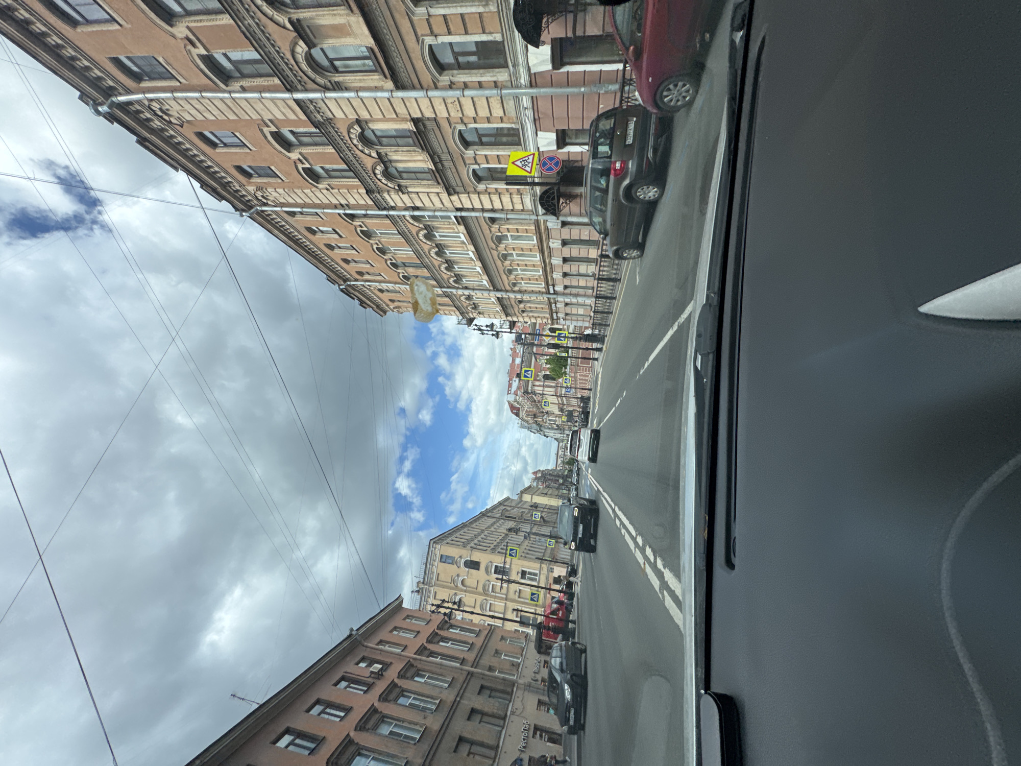 A street scene in Saint Petersburg, Russia, viewed from inside a car. The street is lined with buildings of classical European architectural style, their facades a mix of brown, tan, and beige hues. The sky above is overcast with a thick layer of grey clouds, allowing patches of blue to peek through. The road is clearly marked with white lane dividers, and a few cars are traveling in the same direction as the camera. Street signs are visible at intervals along the road. A pedestrian crossing sign is visible.
