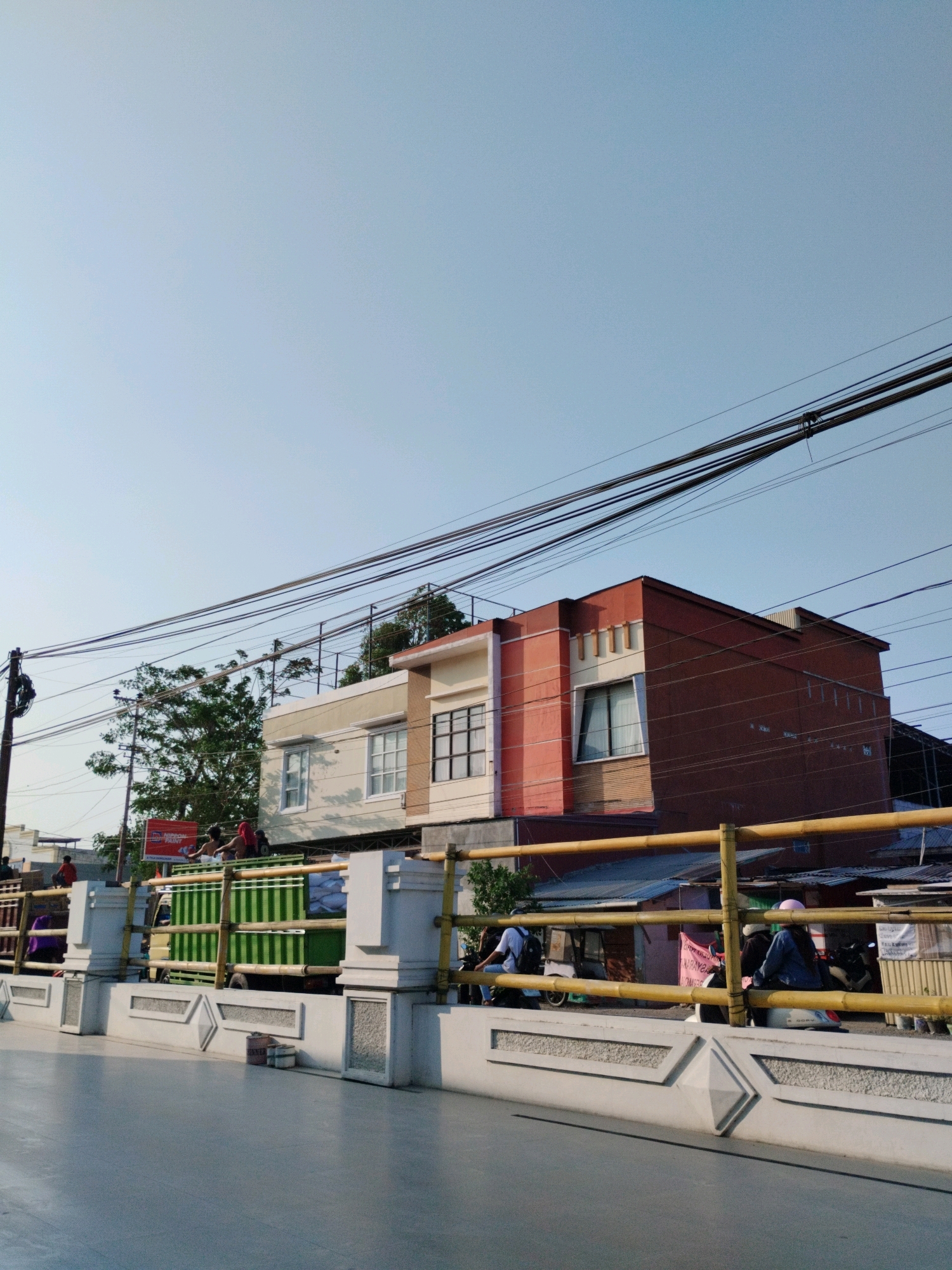 A street-level view of a road in Gorontalo, Indonesia, captures a cluster of buildings and activity under a clear sky. A light-colored concrete barrier with decorative geometric patterns lines the foreground, reflecting the ambient light onto the gray asphalt road. Beyond the barrier, a bright yellow bamboo railing separates the sidewalk from the street.
Several people are visible: three individuals are standing on the back of a green truck filled with white sacks. To the right, near the yellow railing, a person wearing a pink hat sits on a motorcycle. Further down the street, another person is seated on a motorcycle, and a person with a backpack is sitting on the railing.
The buildings in the background exhibit varying architectural styles. One building features a striking orange facade with cream-colored accents around the windows and along the top. Adjacent to it is a lighter-colored building with prominent windows. Trees are interspersed between the structures, adding a touch of greenery. Overhead, a network of black electrical wires crisscrosses the sky, a common sight in urban Indonesian landscapes. The overall impression is a typical urban scene in Gorontalo, showcasing daily life and infrastructure.