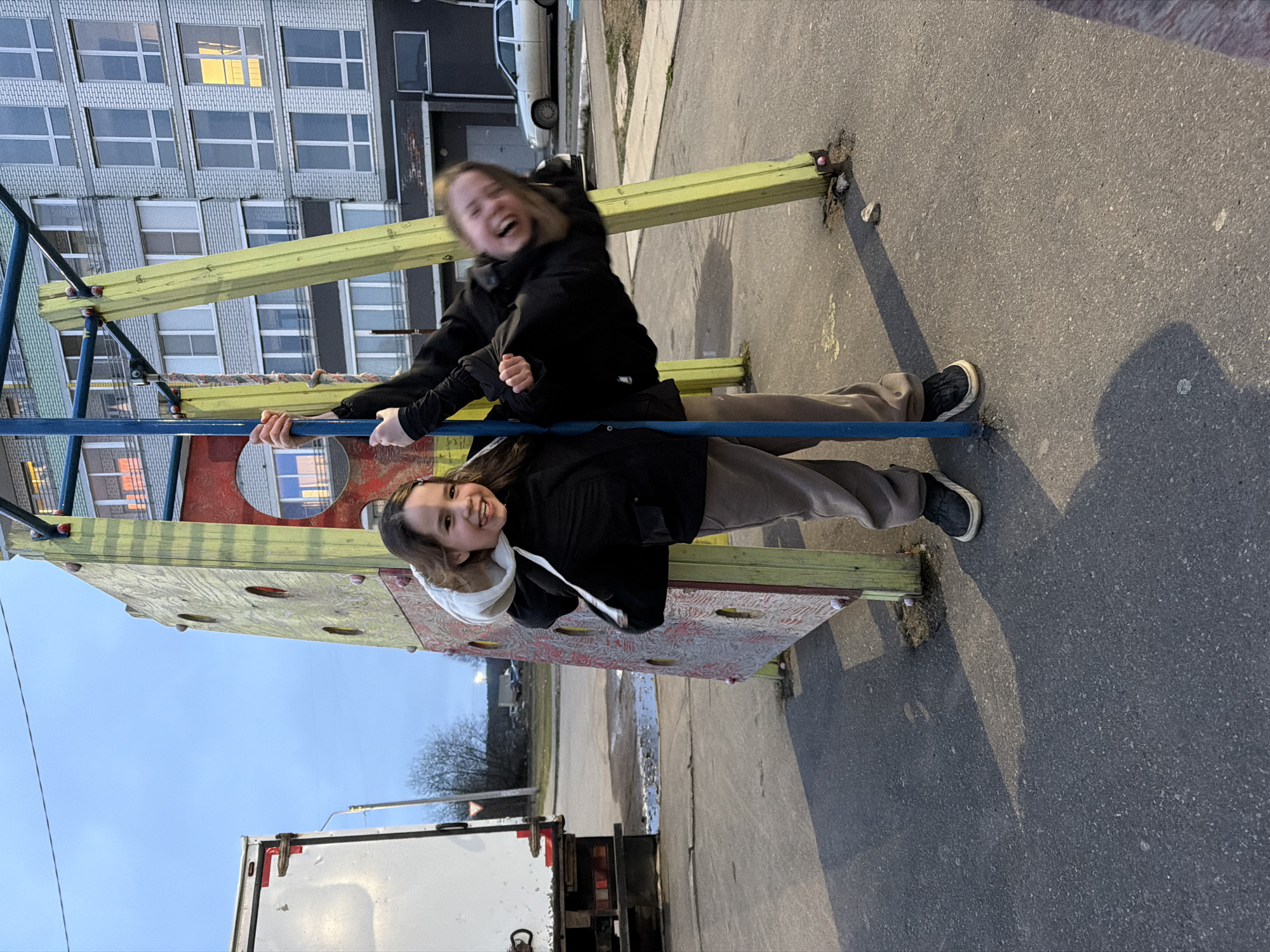 The original image is rotated on its side. When viewed correctly (rotated 90 degrees clockwise), it depicts two young individuals, likely teenagers or young adults, engaged in playful activity on playground equipment in Chaikovsky, Russia.
In the foreground, one girl is smiling directly at the camera. She wears a black jacket with a light-colored hood, dark pants, and black sneakers, and is holding onto a blue horizontal metal bar. Behind and slightly above her, another girl is captured in motion, laughing heartily with her head thrown back, her face blurred due to movement. She also wears a black jacket and dark pants and appears to be hanging from or swinging on a yellow-painted beam or structure.
The playground features a yellow wooden beam, the blue metal bar, and a red and beige climbing wall with various handholds. The ground is asphalt, marked with distinct, elongated shadows, indicating a late afternoon or early evening setting. The sky is clear and a pale blue.
In the background, a large, multi-story residential apartment building with numerous windows is visible, characteristic of a post-Soviet urban environment. One window on an upper floor of the building is illuminated. A white car is partially visible in the mid-ground, and a white truck or van is seen in the lower left section of the frame. The overall atmosphere of the image is joyful and energetic. There is no visible text.