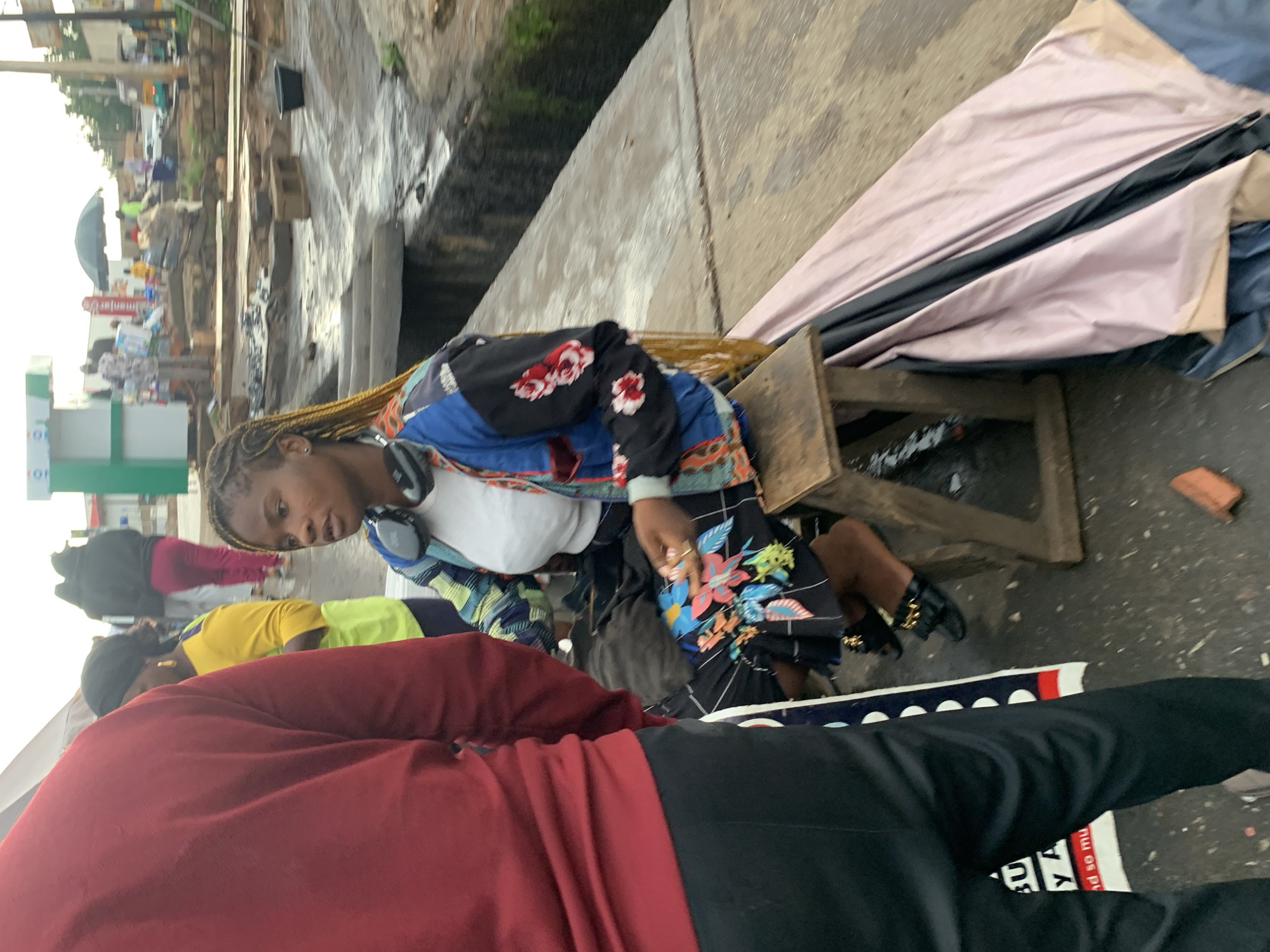 The image depicts a street scene in Barakin Lerawi, Nigeria. A young woman with braided hair and headphones around her neck is seated on a wooden stool, wearing a colorful jacket with floral patterns and a white shirt. She is looking to her right, with one hand resting on her lap. In the foreground, the back of a person in a red shirt and black pants is visible. The background shows a busy street with various people, stalls, and some construction or debris. The ground appears wet, suggesting recent rain.