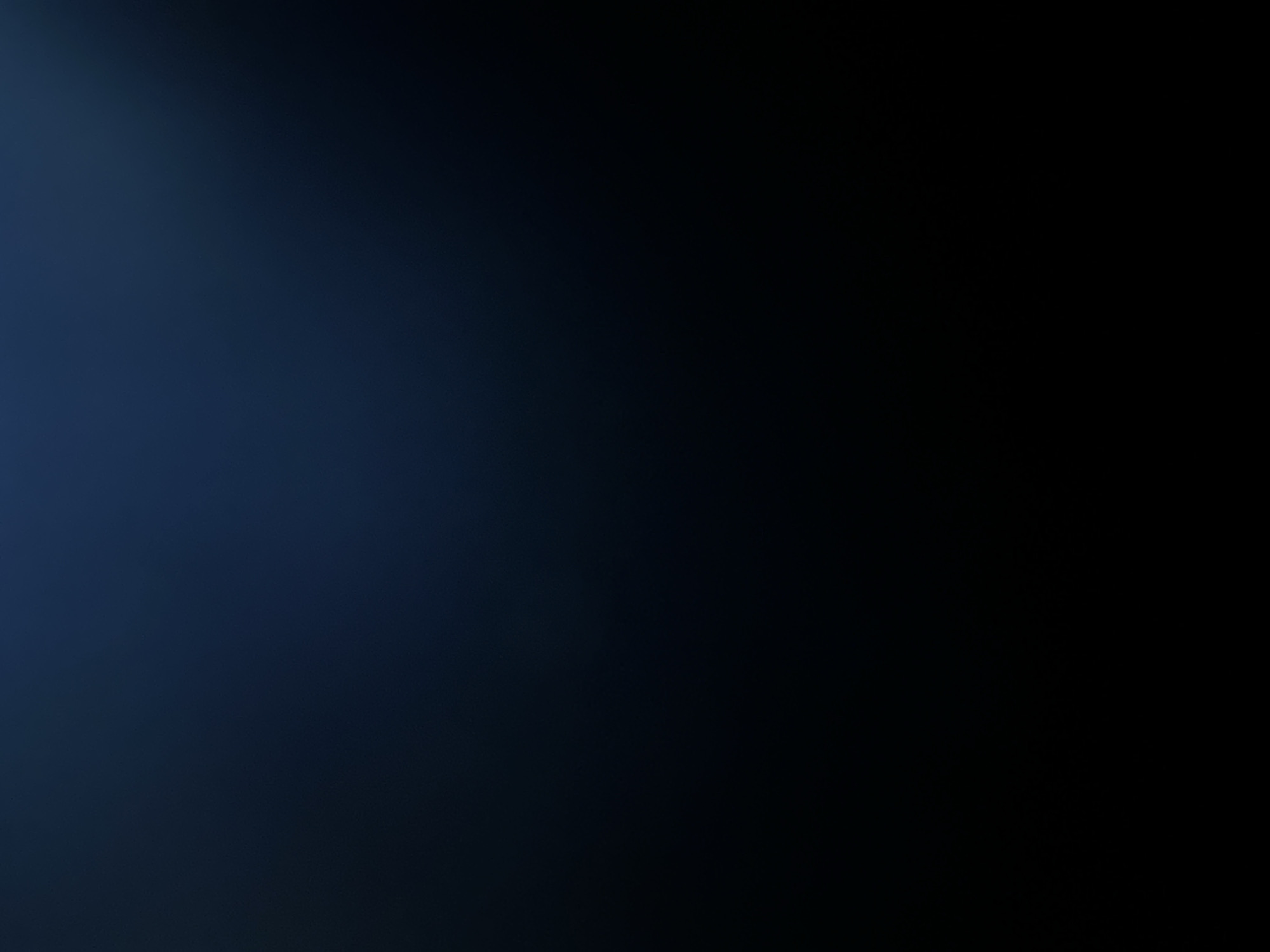 The image is a dark, abstract background with a subtle gradient. The left side of the image is a deep, muted blue, transitioning into a near-black on the right. There is a soft, diffused light source that appears to be coming from the upper left, creating a slight highlight on that portion of the frame. The texture of the background is smooth and matte, with no discernible objects, people, or specific scene elements. The overall impression is one of depth and mystery due to the low light and abstract nature of the image. There is no visible text.