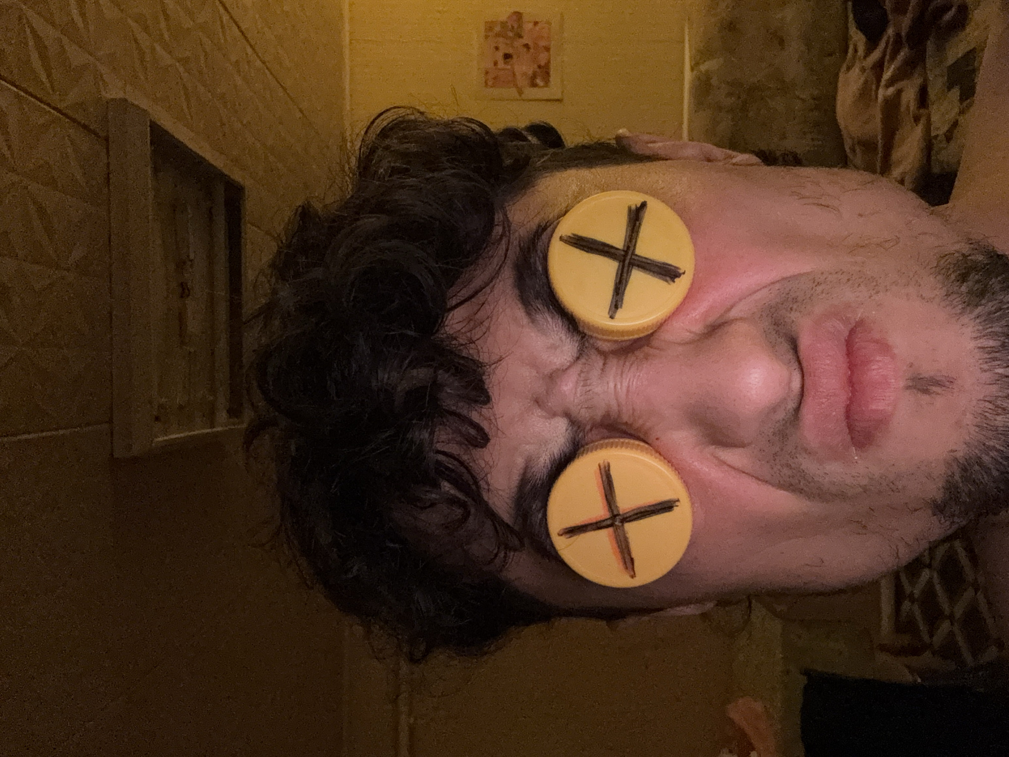 A close-up, front-facing, waist-up shot captures a person's face with two yellow bottle caps, each marked with a black 'X', covering their eyes. The person has dark, curly hair and is making a grimacing facial expression, with furrowed brows and pursed lips. The background is dimly lit and appears to be an indoor setting, possibly a bedroom or living room, with a textured ceiling and a couch visible. The overall lighting is warm and yellow, with some areas in shadow.