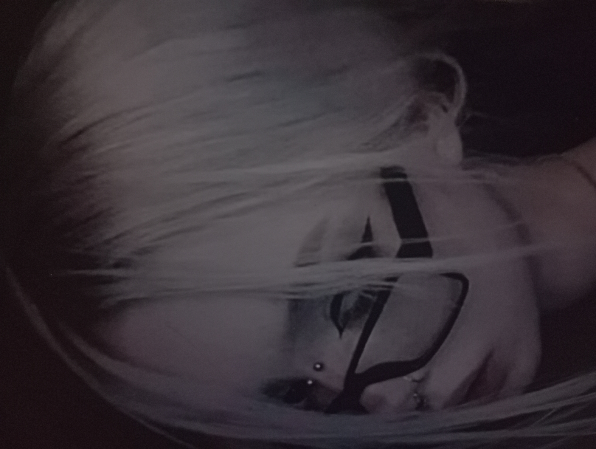 A black and white, high-angle shot depicts a person with pale skin and light, possibly blonde, hair lying on their side. They are wearing black-framed glasses and have multiple piercings on their nose and possibly elsewhere on their face. The background is dark and out of focus, providing no clues as to the location in Odesa, Ukraine.