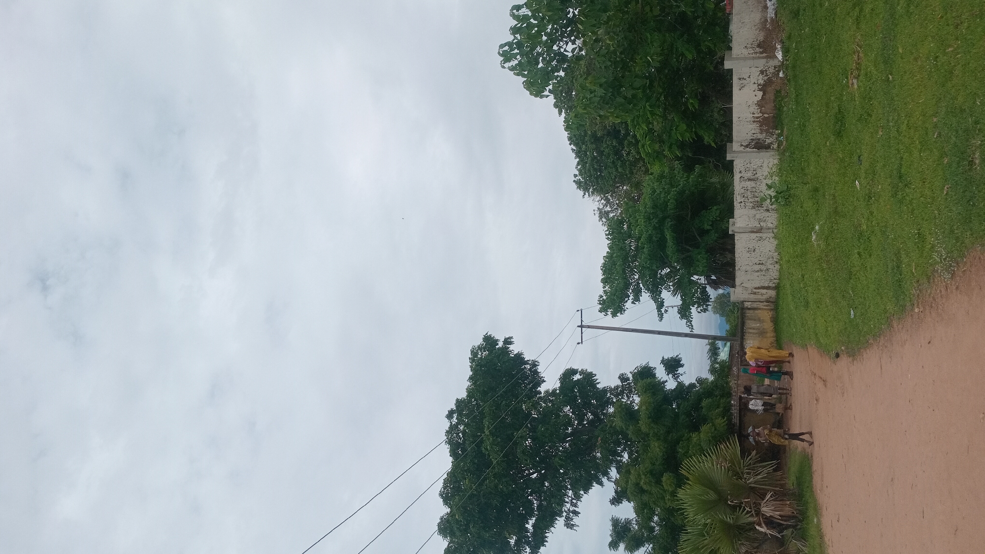 The image depicts a group of people standing on a dirt path next to a grassy area and a concrete wall. Several trees with green foliage are visible in the background, as well as utility poles and wires. The sky is overcast, suggesting a cloudy or possibly rainy day. The people appear to be casually gathered, with no obvious signs of distress or specific activity. Their clothing suggests a warm climate. The overall setting appears to be a rural or semi-rural area, possibly a village or town outskirts. The time of day is unclear due to the overcast sky, but it appears to be daytime. There is no visible text in the image.