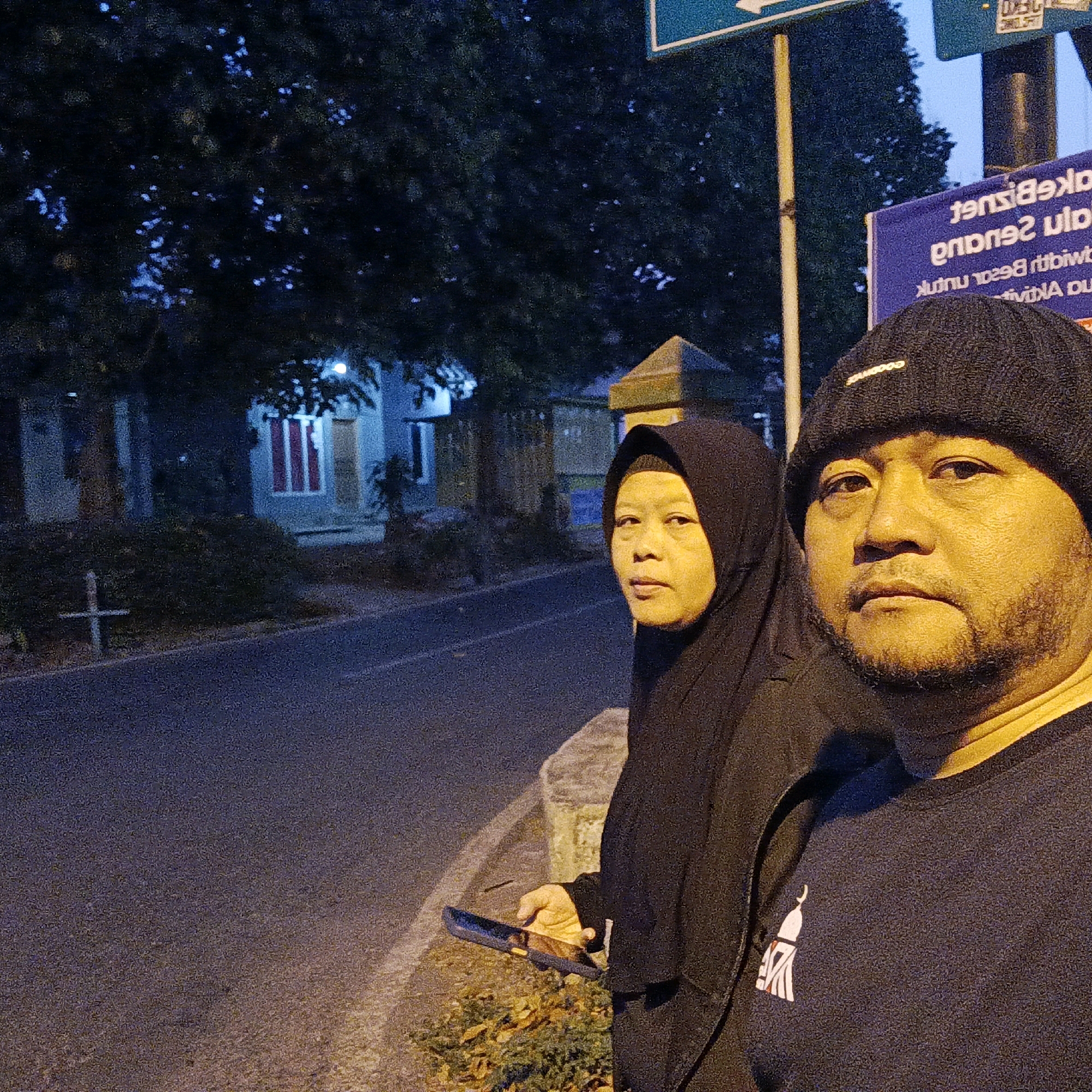 Two individuals, a man and a woman, are standing on the side of a road at dusk or night in Salatiga, Indonesia. The man is in the foreground, facing the camera and wearing a black beanie and a dark t-shirt with a white emblem. The woman is behind him and to his left, wearing a black hijab and a black garment. She is looking towards the camera. The background shows a street, trees, and a building with illuminated windows. There is also a signpost with text visible. The man is holding a smartphone in his left hand. The overall lighting suggests it is evening, possibly under artificial streetlights.
