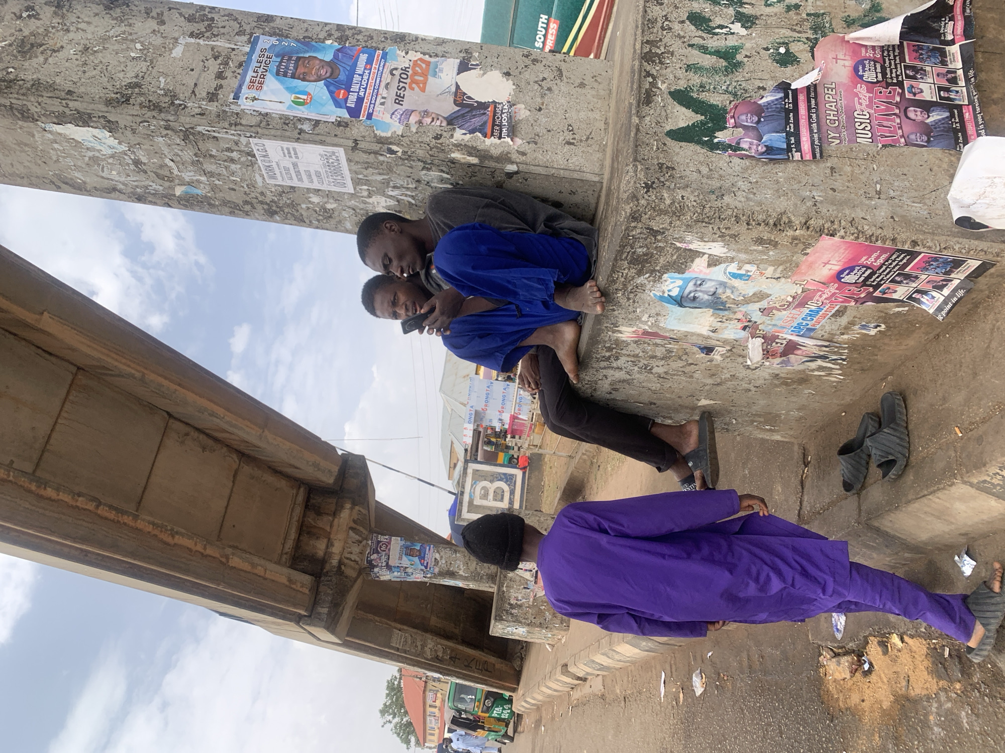 Here's a forensic description of the image:
**Location:** Jos, Nigeria. Evidenced by "Jos South Constituency" text on a political poster affixed to a concrete column.
**People:** Three males are visible. Two younger males are sitting on a concrete shelf, likely the base of a column or bridge support, looking at a mobile phone. One is wearing blue pants and a gray shirt; the other is wearing blue pants. The third male is walking on the sidewalk wearing a purple robe and a black cap. He is moving away from the camera's perspective.
**Objects:** The immediate environment includes a concrete column covered in posters, some torn. Posters advertise political campaigns, music events ("NY Chapel," "Music Time Live"), and job vacancies ("WORK@N.G.O"). Under the concrete shelf is littered with used slippers. The background contains buildings and vehicles, indicating an urban setting. A bridge or overpass is visible overhead.
**Actions/Interactions:** Two males are engaged with a mobile phone. The third male is walking. The sitting males seem to be occupying the space around the column as a seating area. The presence of numerous posters suggests active posting of information in this public space.
**Environment:** The environment appears to be an urban area with pedestrian traffic near a road, possibly a bus stop or gathering point, as indicated by the number of people. The air appears hazy/dusty.