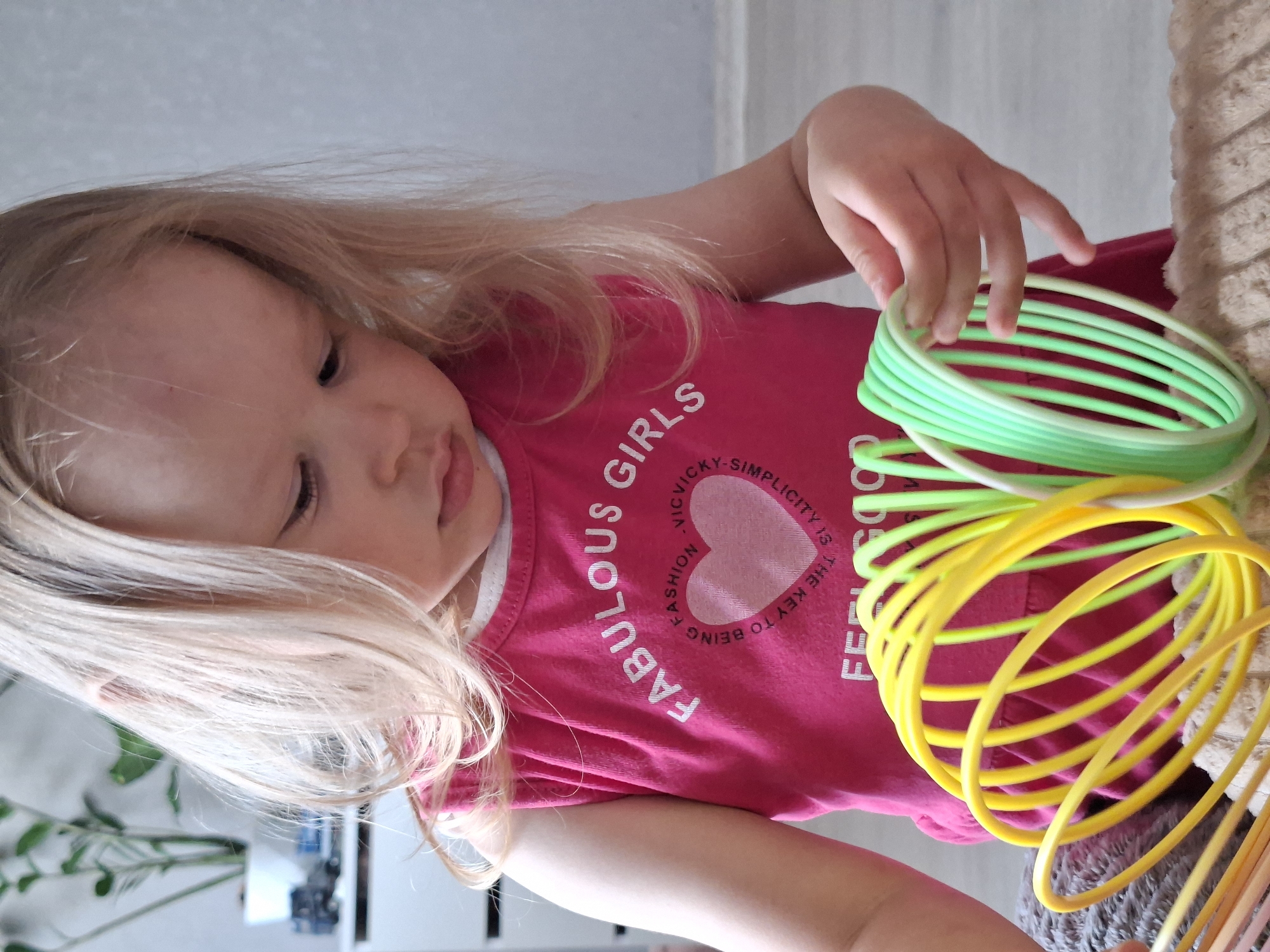 A young girl with blonde hair is playing with colorful plastic coil bracelets. She is wearing a pink t-shirt with the word "Fabulous" printed on it. The scene appears to be indoors. The location and city are unknown.
