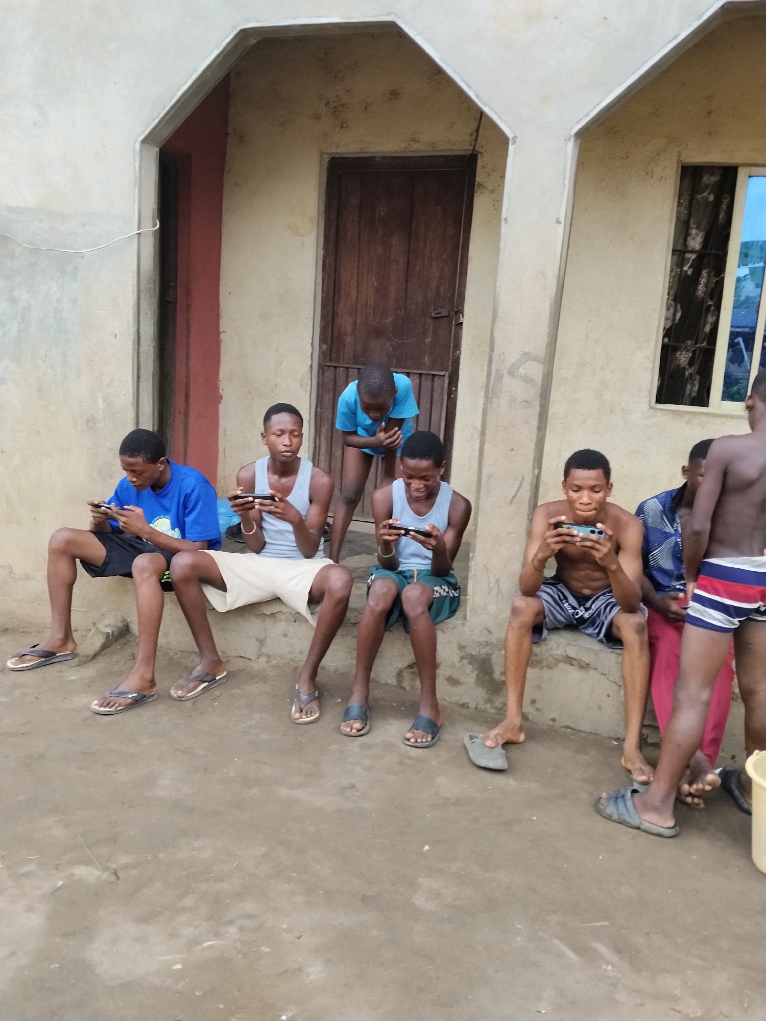 A group of approximately seven young men are gathered outdoors in Aba, Nigeria. They are positioned on or near the steps of a building with a weathered exterior. Four of the individuals are seated and are actively engaged with their mobile phones, holding them in landscape orientation. One individual is standing and leaning over to observe another's phone. Several of the men are wearing casual clothing such as shorts, tank tops, and t-shirts. The men all have bare feet or are wearing flip-flops. The building features a wooden door and a small window with a visible curtain. The ground is a concrete or paved surface.