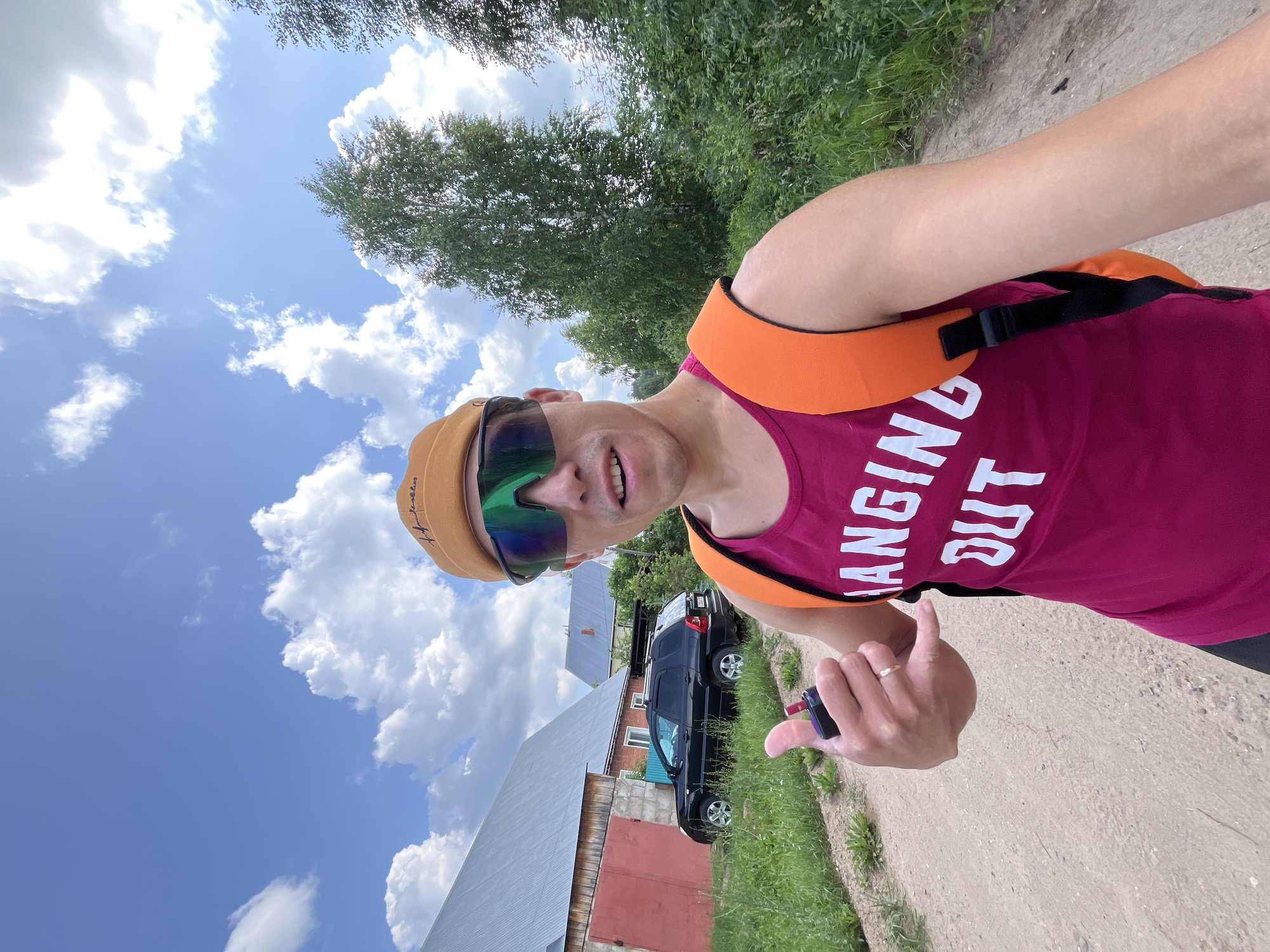 A man wearing a pink tank top with white lettering that reads "HANGING OUT", an orange backpack, and a tan baseball cap with a logo is posing for a selfie. He is wearing large, mirrored sunglasses and making a "shaka" hand gesture while holding a dark-colored object, possibly a vape pen. The man is standing on a dirt road in front of a rural house with a red garage door and a black SUV parked in the driveway. The background features green trees and a blue sky with white clouds. The scene appears to be in the city of Chaikovsky, Russia, given the architectural style of the house and the surrounding vegetation.