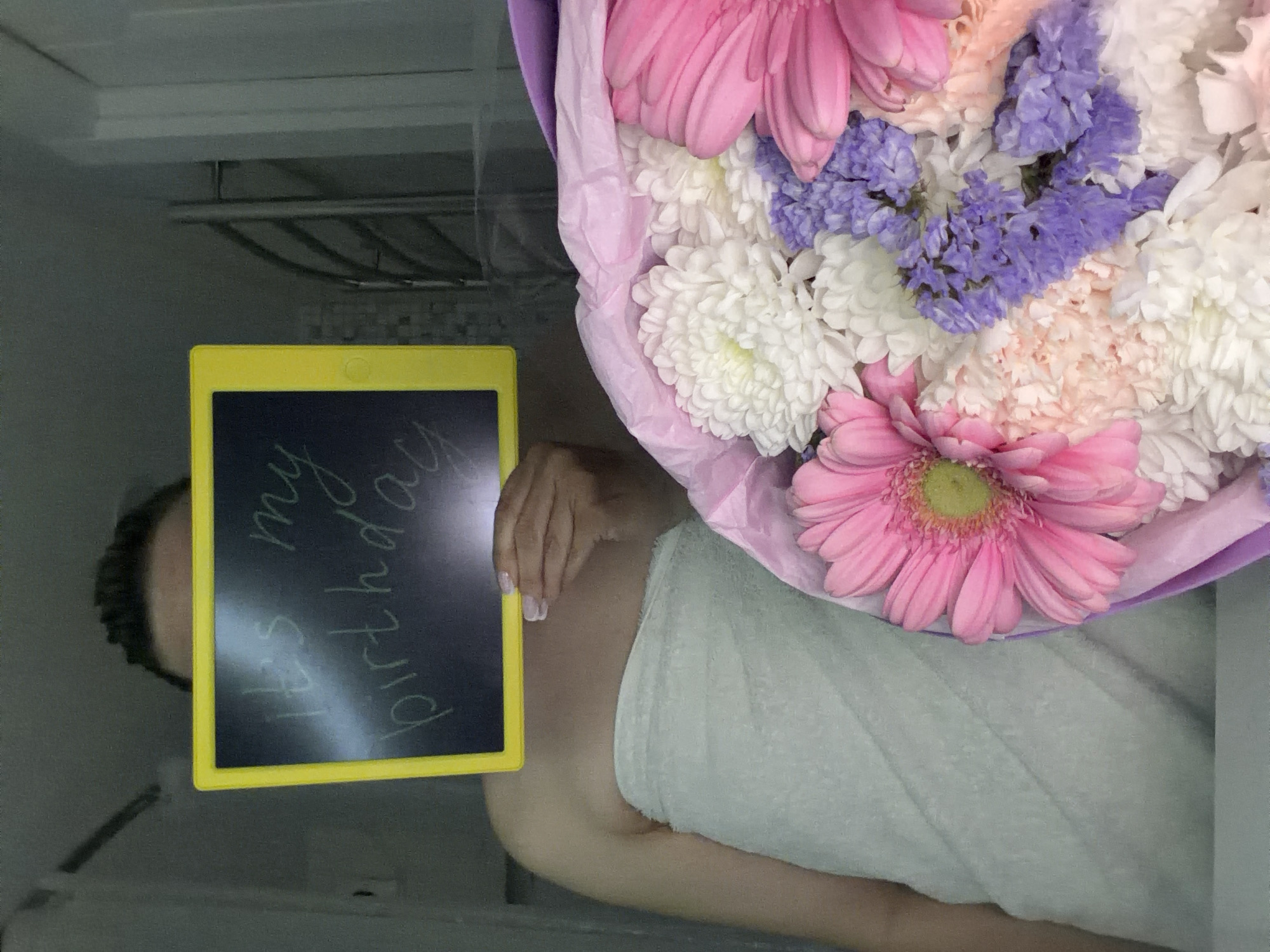 A person wrapped in a light green towel holds a yellow-framed electronic writing board displaying the words "It's my birthday." A bouquet of pink, white, and purple flowers wrapped in pink and purple paper is positioned next to the person. The background appears to be the interior of a refrigerator. No specific information suggests this is located in Votkinsk, Russia.