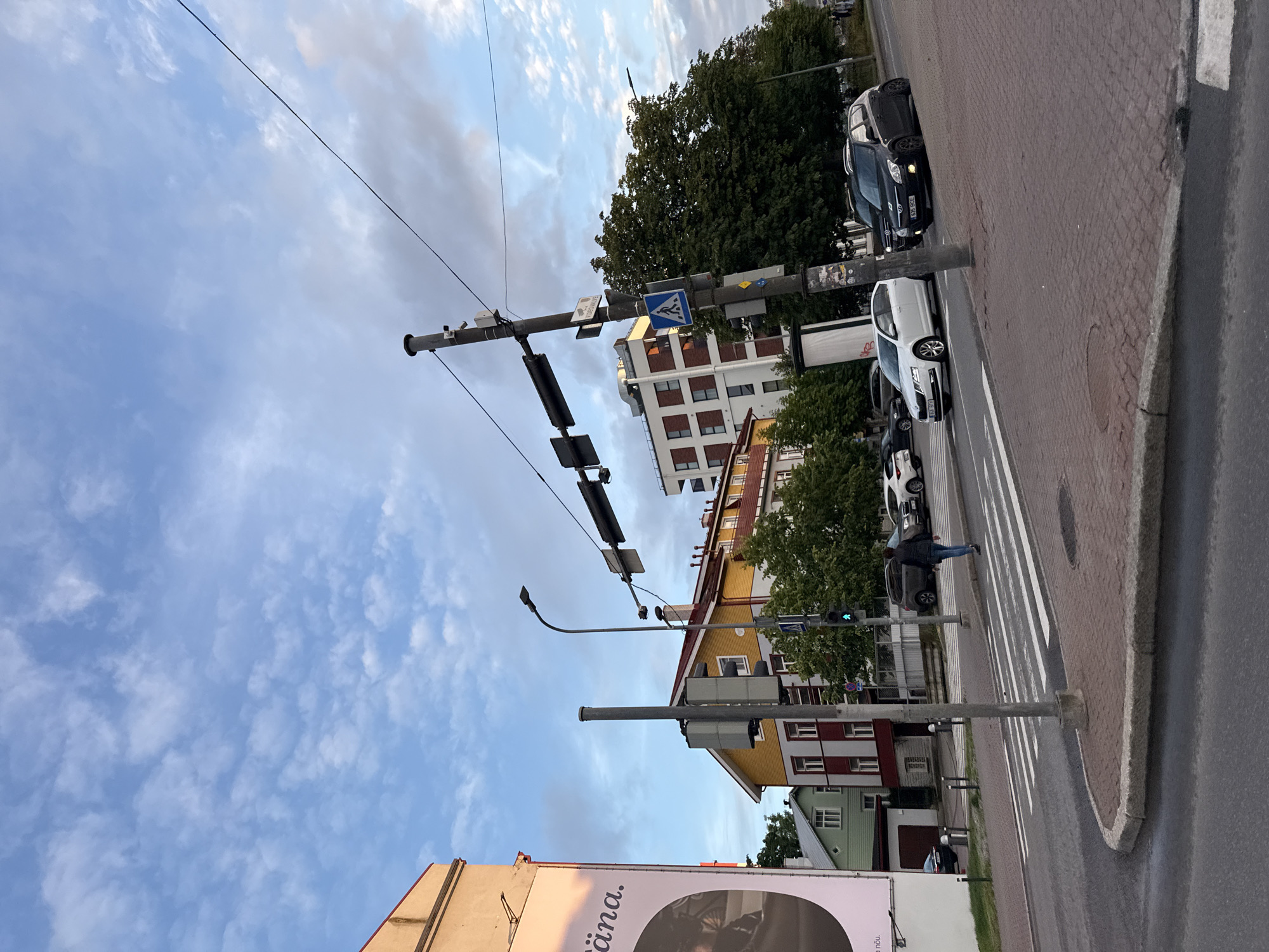 The image captures a street scene in Tallinn, Estonia, likely during twilight or a cloudy day, given the soft lighting and scattered clouds in the sky. In the foreground, a pedestrian crossing with white stripes is visible on a paved road, with red brick paving on the sidewalk. Several cars are present: a dark sedan, a black hatchback with license plate "635 BCE", and a white sedan.
Dominating the midground are several buildings of varying architectural styles and colors. To the left, a tall building with a pale facade features a large billboard with text in an unknown language. Adjacent to it is a two-story building painted green, followed by a multi-story building with a bright yellow facade and a red roof, featuring multiple windows and balconies. Further in the background, a modern white building with red-brick accents rises, interspersed with dense green trees.
A traffic light pole with traffic signals and a pedestrian crossing sign is positioned prominently. A person is seen walking across the pedestrian crossing from left to right. The scene is completed by overhead wires and streetlights, typical of an urban environment.