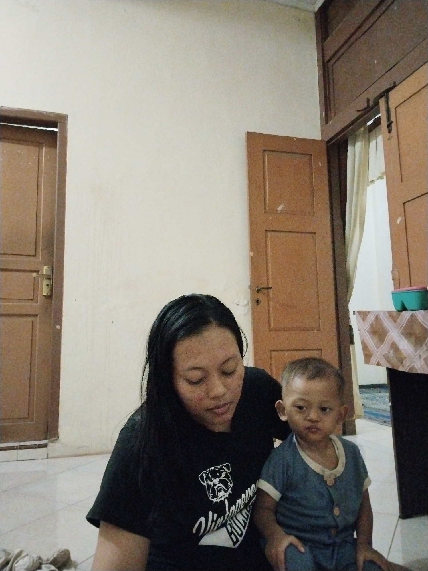 In this image, a woman and a young child are seated on a tiled floor. The woman has long, dark hair and is wearing a black t-shirt with a graphic of a dog. The child, with short, light-colored hair, is dressed in a blue collared shirt and pants. They appear to be indoors, as suggested by the wooden doors and wall in the background. To the left, a wooden door with a brass handle is visible, and to the right, another wooden door is ajar, revealing a light-colored curtain. A small table with a patterned cloth and a turquoise container is also present on the right side of the frame. The scene has a casual, indoor setting, possibly a home. The lighting suggests it is daytime. The overall mood is calm and domestic. The text on the woman's shirt is partially visible and appears to say "VINTAGE BULL...".