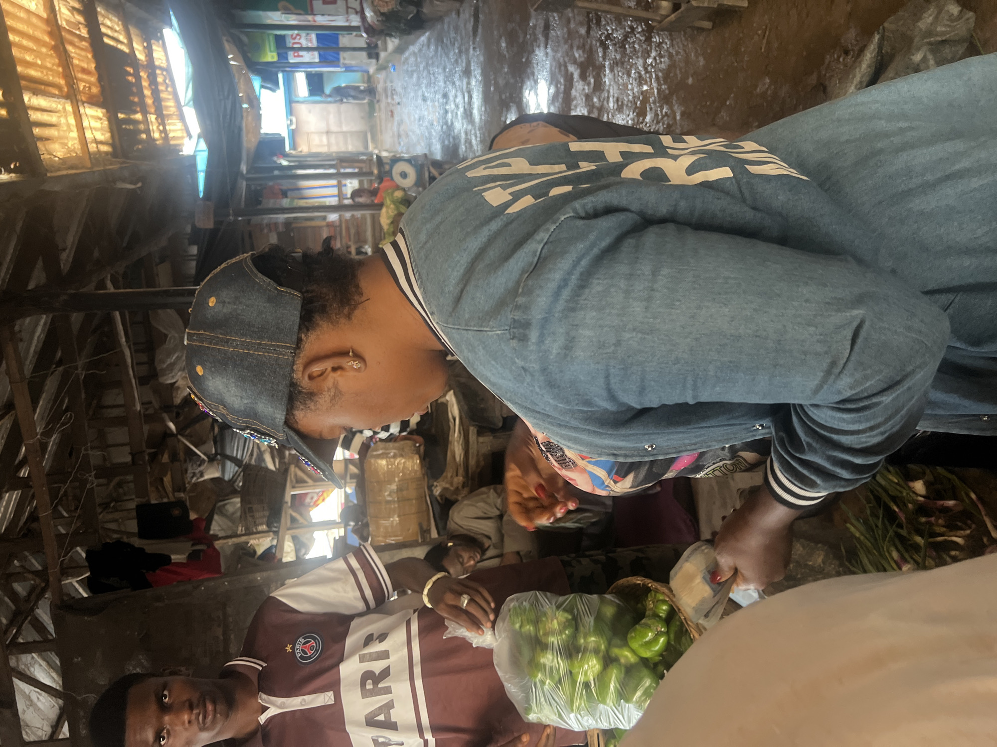 The image depicts a vibrant marketplace scene in Oko Amakom, Nigeria, likely during daylight hours, given the ample natural light filtering through the corrugated metal roofing. In the foreground, a man wearing a maroon and white polo shirt with "PARIS" emblazoned across it is standing to the left. He has a ring on his left hand and is holding a clear plastic bag filled with green bell peppers. To his right, a woman wearing a denim jacket and a baseball cap is partially visible, facing away from the camera. She is holding a wad of cash in her hands, suggesting a transaction is taking place. Behind them, other vendors and shoppers can be seen, contributing to the bustling atmosphere of the market. The ground appears to be wet and muddy, common in outdoor markets after rain. The market stalls are simple, constructed with wood and metal, and laden with various goods. The overall impression is one of a lively and active commercial hub within the community.