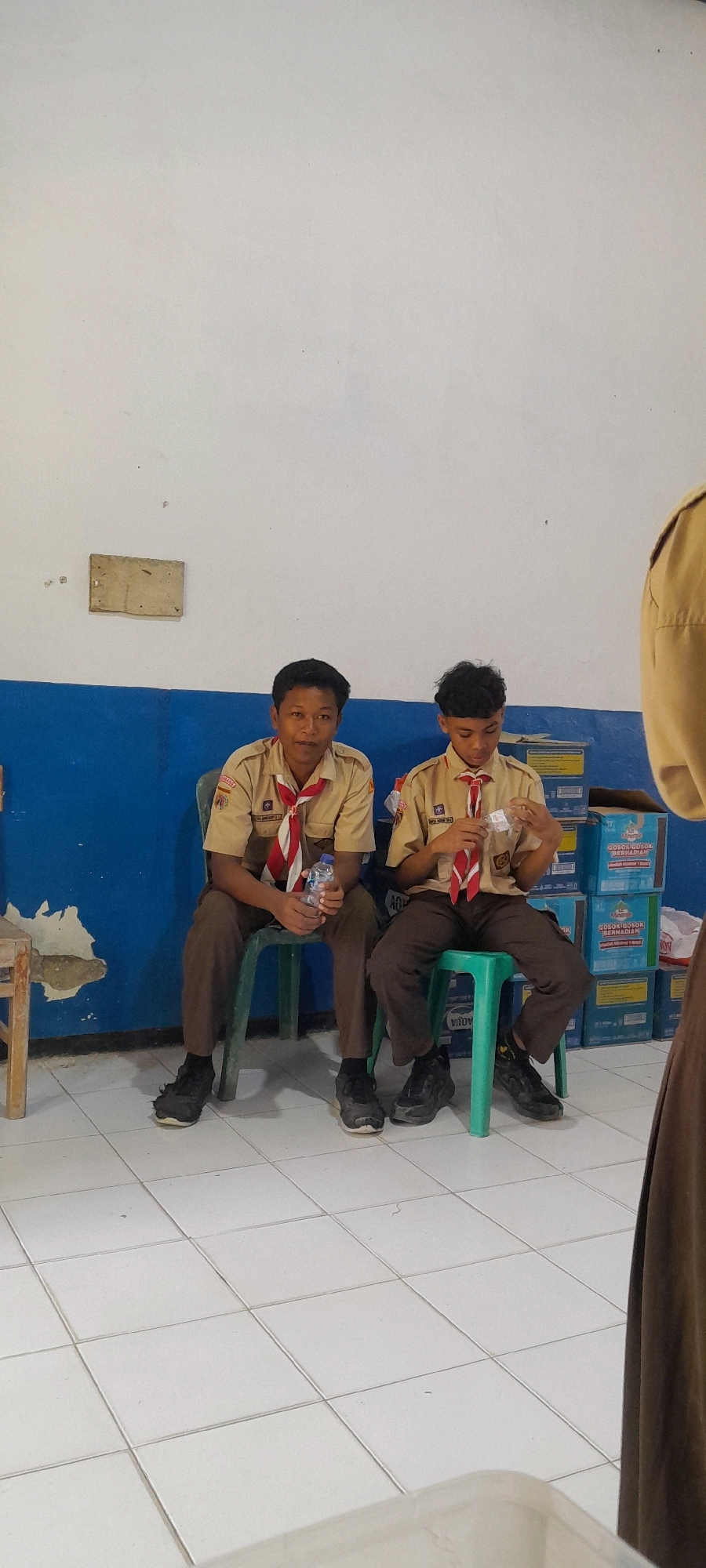 The image shows two young boys in scout uniforms sitting on chairs in front of a white wall with a blue stripe. The boy on the left is wearing a tan uniform shirt with a red and white scarf and brown pants, and is holding a water bottle. The boy on the right is wearing a similar uniform and is holding a small object in his hands. In the background, there are stacks of boxes. The floor is tiled with white squares. The lighting suggests an indoor setting, possibly a community hall or classroom in Tahunan, Indonesia, given the context provided.