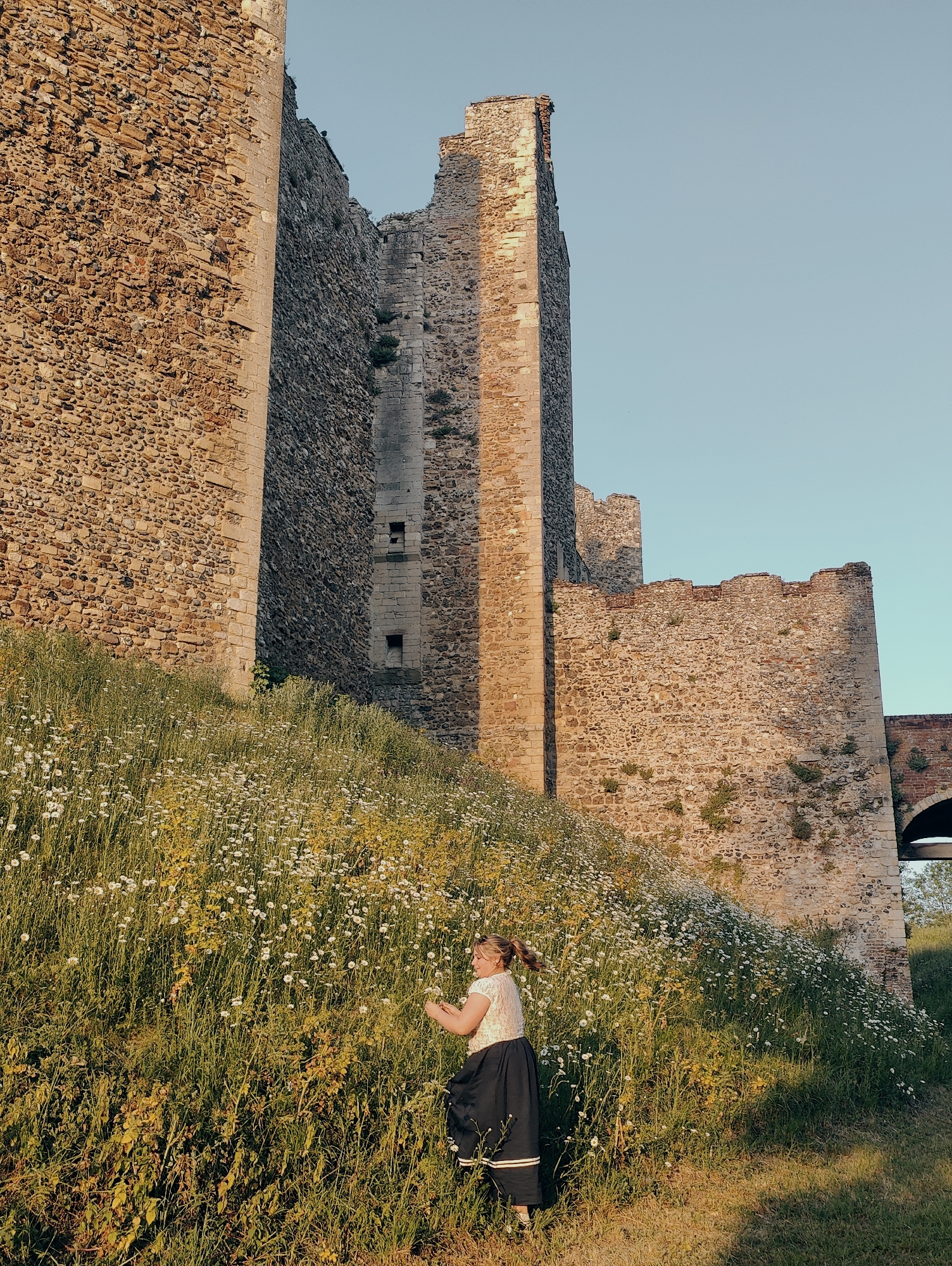 A young woman stands on a grassy, wildflower-covered hillside near Framlingham Castle in East Suffolk, United Kingdom. The castle's stone walls rise in the background, lit by the late afternoon sun. The woman is wearing a white blouse and a long, dark skirt, and she appears to be picking flowers.
