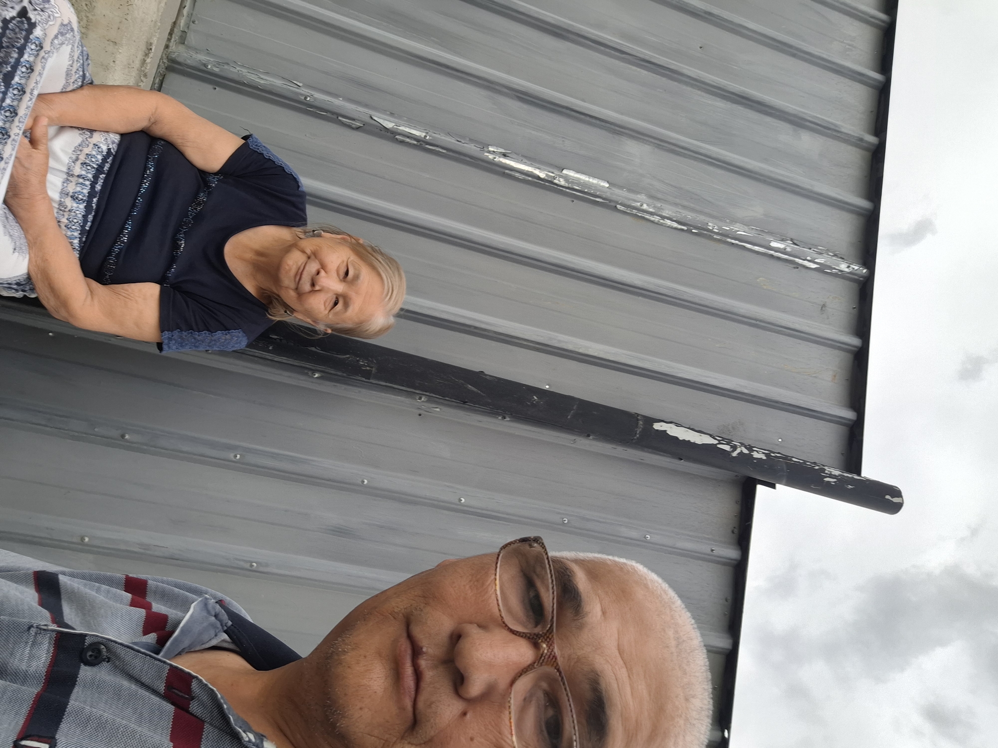 In Almaty, Kazakhstan, an outdoor selfie is taken with two subjects against a corrugated metal wall. The woman, seated on the left, is wearing a dark blue shirt with blue lace accents on the sleeves and a patterned skirt. She has fair skin and light brown hair, and is looking at the camera with a slight smile. To her right, a man is taking the selfie, his face in the foreground, wearing glasses and a striped polo shirt. He is looking directly at the camera. A dark, weathered pole stands between the two subjects, with peeling paint. The background is a gray, corrugated metal wall and a cloudy sky.