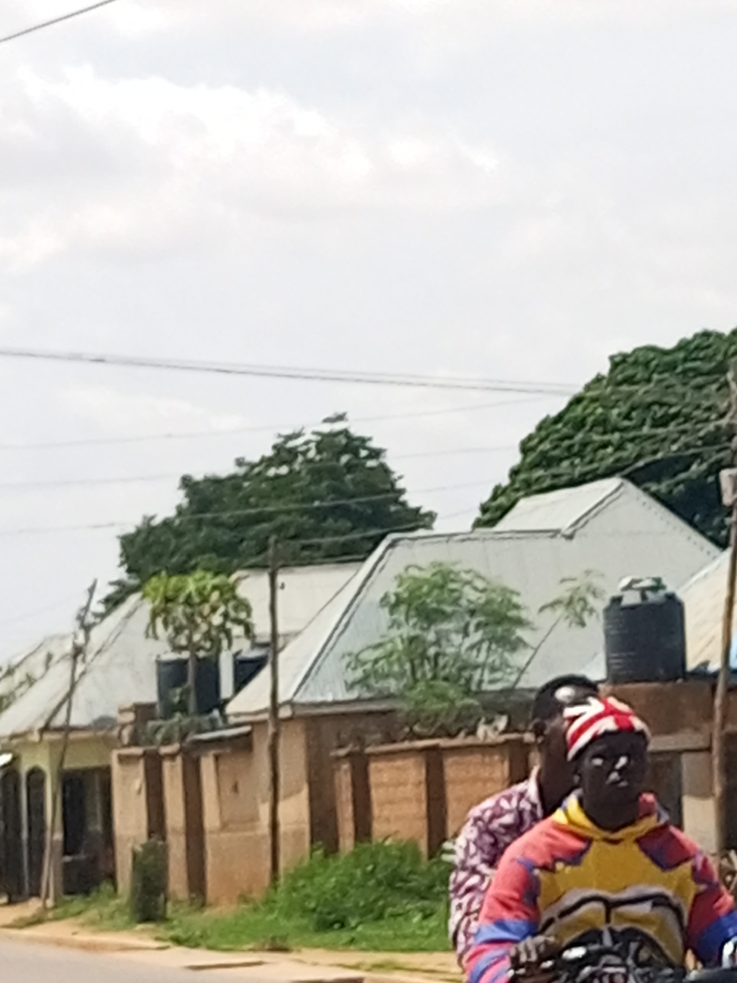 The image shows a street scene in Tempure, Nigeria, with several residential buildings lining the road. The buildings have corrugated metal roofs and are of a simple construction, with some appearing to be under renovation or in disrepair. There are water tanks visible on some of the rooftops. Lush green trees and vegetation are present, indicating a warm climate. In the foreground, two individuals are riding on a motorcycle. The person in front is wearing a colorful hooded sweatshirt and a hat with a pattern resembling the Union Jack. The person behind them is wearing a patterned shirt. The scene is captured during daylight, with an overcast sky.