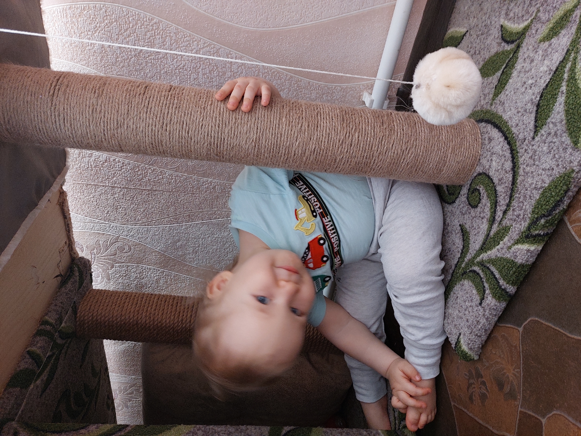 A young child with blond hair, wearing a light blue shirt with a car graphic and gray pants, is holding onto a scratching post covered in brown rope. The post has a white fuzzy ball attached to it. The child is upside down, looking at the camera, with the post appearing above them. The background includes a patterned wallpaper, part of a cat tree, and a rug with a green leafy pattern. The photo appears to be taken indoors. The location is Большой Букор, Russia.