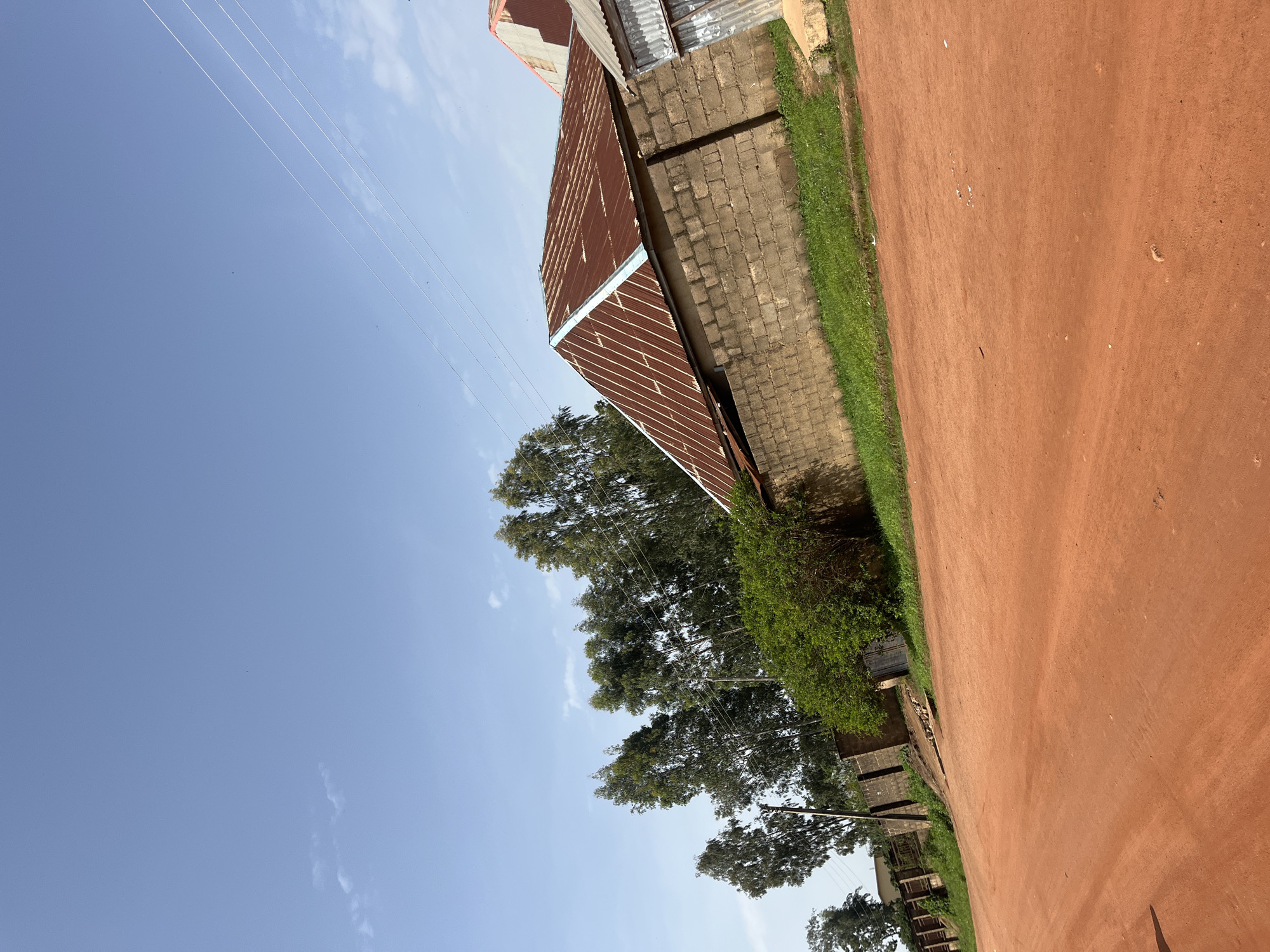 In Bukuru, Nigeria, a dirt road with a reddish-orange hue occupies the foreground, angled from the bottom left to the middle right of the frame. Along the right side of the road, a low wall constructed of grey cinder blocks runs horizontally, topped by a lush strip of green grass. Behind the wall, a building with a rusty, corrugated metal roof and similar cinder block construction is visible, with a second building's roof visible further back. Tall, slender trees with dense green foliage line the background, extending across the left and middle sections of the image. The sky above is a clear, pale blue, with a few wispy white clouds scattered across the upper left. Electrical wires are strung diagonally across the upper right portion of the sky. The scene appears to be an outdoor view of a residential or semi-rural area.