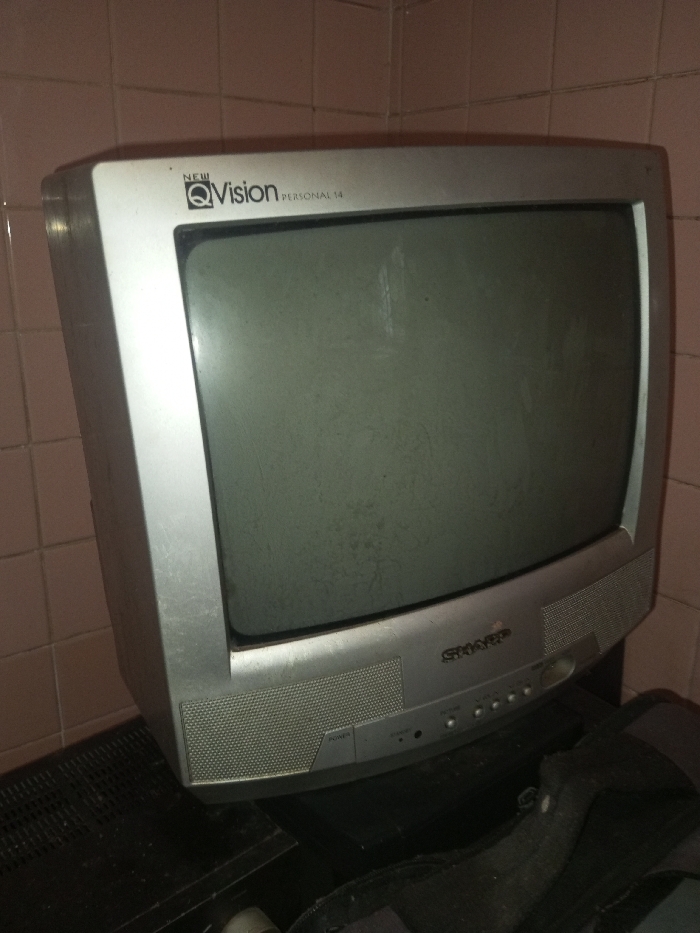 An old, dusty silver CRT television rests on a dark surface. The television, labeled "New Vision Personal 14," features a rectangular screen and a lower panel with speaker vents and several small, round buttons. The screen appears gray and dirty. The background features a tiled wall with light pink tiles. The overall setting looks worn and dated. A portion of a dark object is visible below the TV.