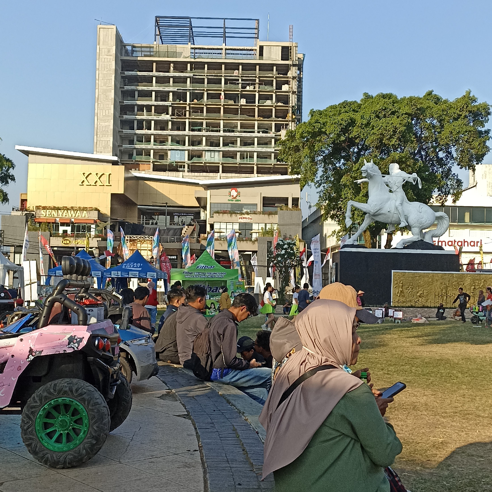 This image captures a street scene in Magelang, Indonesia, with a focus on a large white statue of a man on horseback. Behind the statue, a multi-story building, possibly under construction or renovation, is visible against a clear blue sky. In the foreground, a pink and blue all-terrain vehicle (ATV) is parked. Several people are present in the scene, some sitting on a low wall and others standing. They are dressed in casual attire. Various colorful tents and flags are set up, suggesting an outdoor event or market. The ground is covered with grass, and the overall lighting indicates it is daytime, likely afternoon, given the long shadows. The specific location in Magelang is identified by the presence of the XXI cinema building, a common landmark in Indonesian cities.