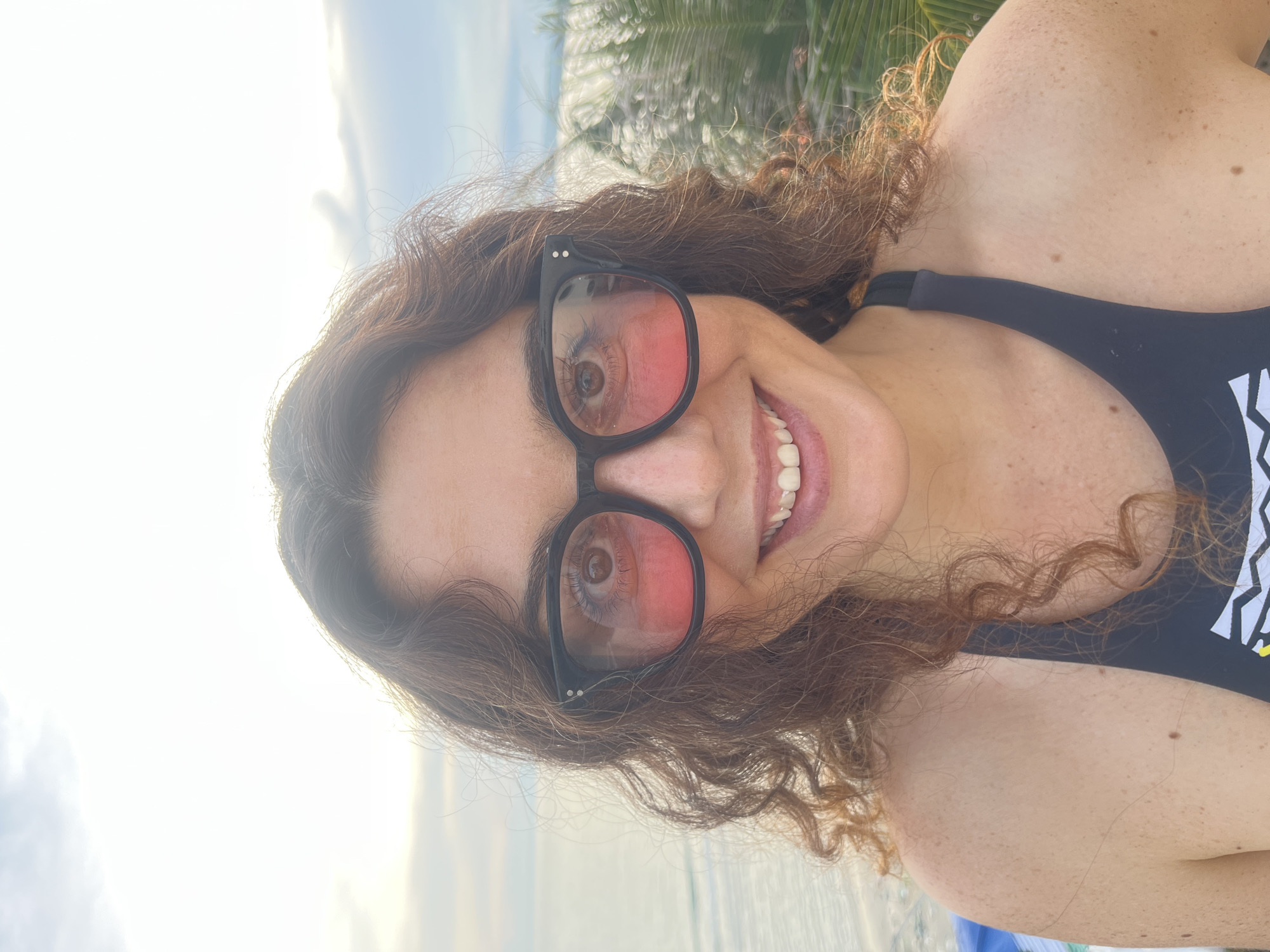 A woman with curly brown hair and freckled skin is posing for a close-up selfie outdoors. She is wearing large, black-framed sunglasses with pink lenses and a black swimsuit. Her eyes are visible through the sunglasses, and she is smiling broadly, revealing her teeth. The background suggests a tropical location with palm trees and the ocean under a cloudy sky, possibly the coast of Mũi Né, Vietnam, known for its beaches and palm-lined shores.
