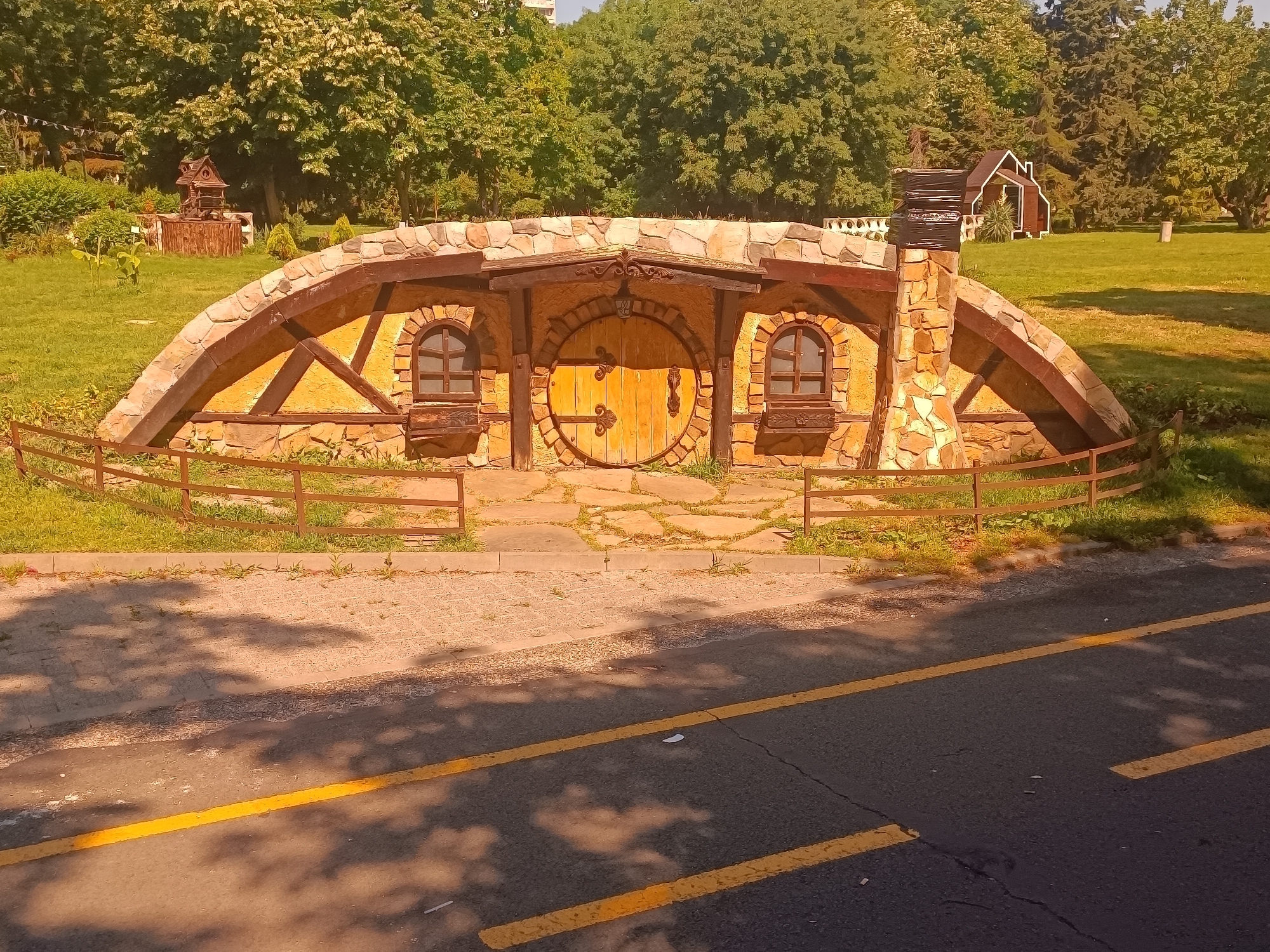 The image depicts a whimsical, hobbit-hole-style dwelling situated in the Sea Garden of Burgas, Bulgaria. The structure is built into a grassy embankment and features a large, round wooden door with ornate metal hinges and handles. Flanking the door are two arched windows with wooden frames and a decorative planter box beneath each. The facade is constructed with stone and has a rustic, charming aesthetic, with exposed wooden beams supporting the roof and a stone chimney on the right. A low, brown metal fence encloses the front of the dwelling, separating it from a paved walkway made of irregularly shaped stones. Beyond the structure, the Sea Garden unfolds with lush green grass, mature trees, and in the distance, a small, stylized wooden house. The foreground shows a section of asphalt road marked with two yellow lines, with dappled sunlight and shadows cast by trees. The scene is bathed in warm, late afternoon light. No people are visible in the image.