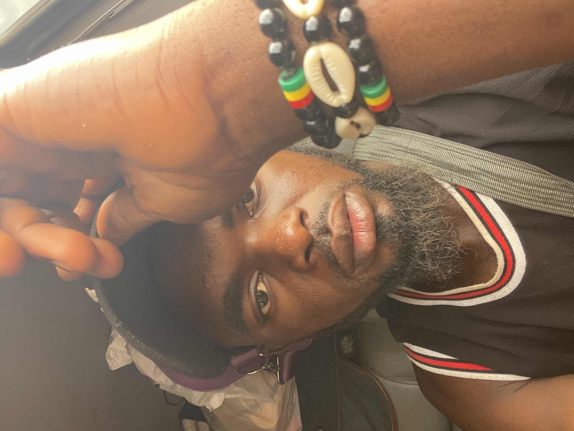 A close-up, low-angle portrait of a dark-skinned man with a salt-and-pepper beard. He is wearing a black t-shirt with red and white trim around the collar and a bracelet made of black beads, cowrie shells, and beads of red, yellow, and green on his wrist. The background is blurry, but suggests he is indoors, possibly in a car based on the angle of the view and the presence of a car roof visible in the upper left corner of the image. The location is Otukpa, Nigeria, but there's no confirmation of this information from the image alone. The man's expression cannot be determined from this image.