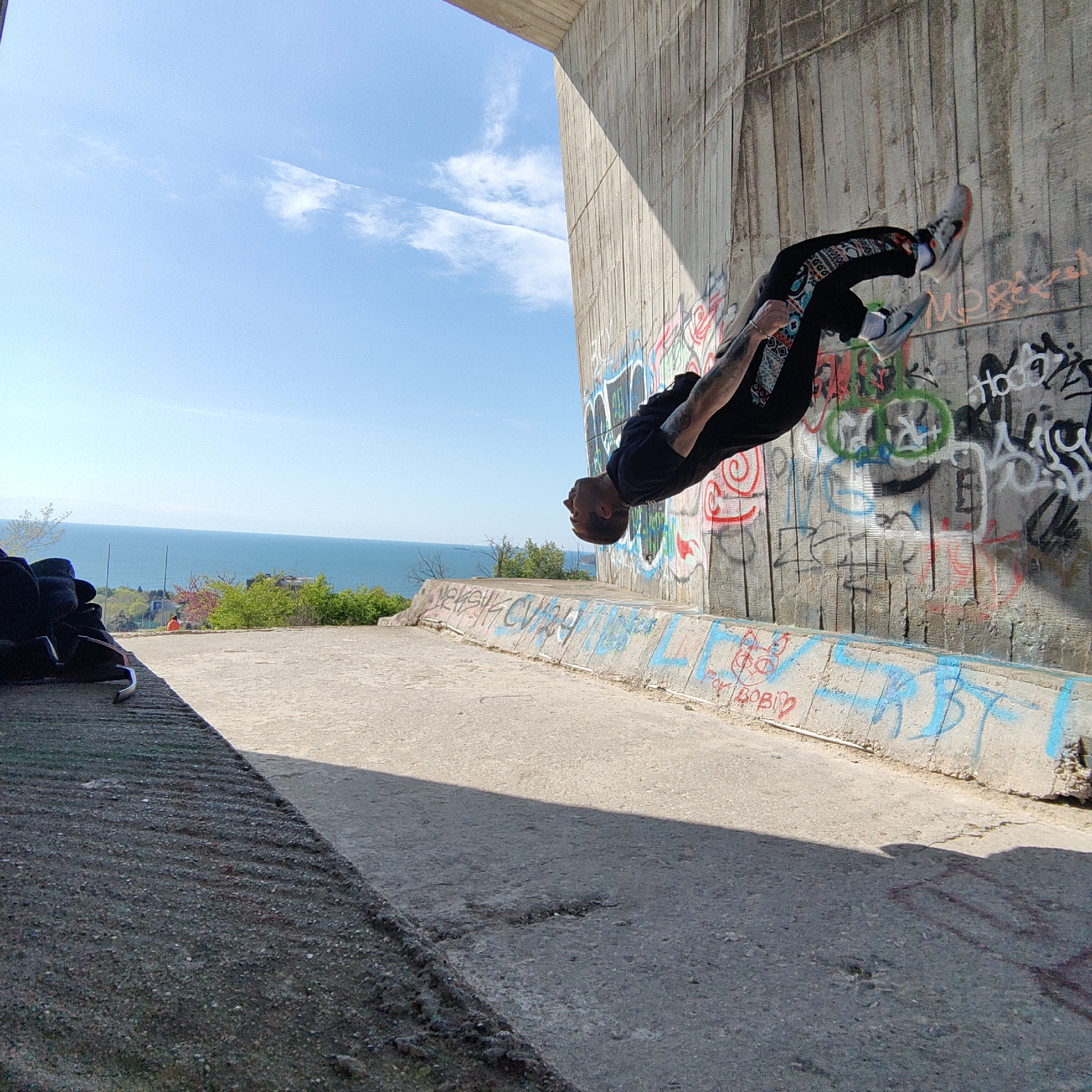 A man is captured mid-backflip against a graffiti-covered concrete wall. The location appears to be under a bridge or overpass, with the blue sky and the Black Sea visible in the background. The image was taken in the city of Varna, Bulgaria.
The man is wearing a black t-shirt, patterned black pants, and sneakers. He has tattoos on his arms. His body is arched backwards, with his head pointing towards the ground and his legs towards the sky.
The concrete wall is covered in various colorful graffiti markings. A low concrete barrier runs along the edge of the platform where the man is performing the flip, also covered in graffiti.
In the background, a glimpse of the Black Sea is visible, as well as some greenery and a distant building. The sky is a clear blue with a few wispy clouds. The ground under the bridge is concrete. A black duffel bag sits near the bottom left corner of the image.
