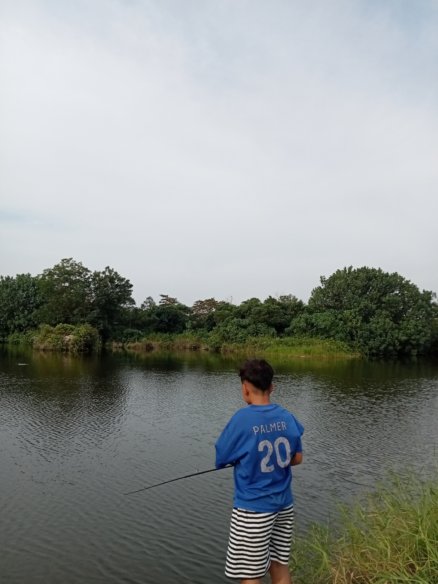 In the city of Kualu, Indonesia, a young boy, viewed from behind, stands by a calm body of water, likely a lake or river, engaged in fishing. He is wearing a blue t-shirt with the name "PALMER" and the number "20" printed on the back, along with black and white striped shorts. The boy holds a fishing rod, its tip extended over the water. The surrounding environment is lush with dense green trees forming a natural backdrop along the far bank of the water. The sky above is overcast with a light, diffused gray. The water's surface is rippled, reflecting the muted light. Tall green grass grows along the bank where the boy is standing.