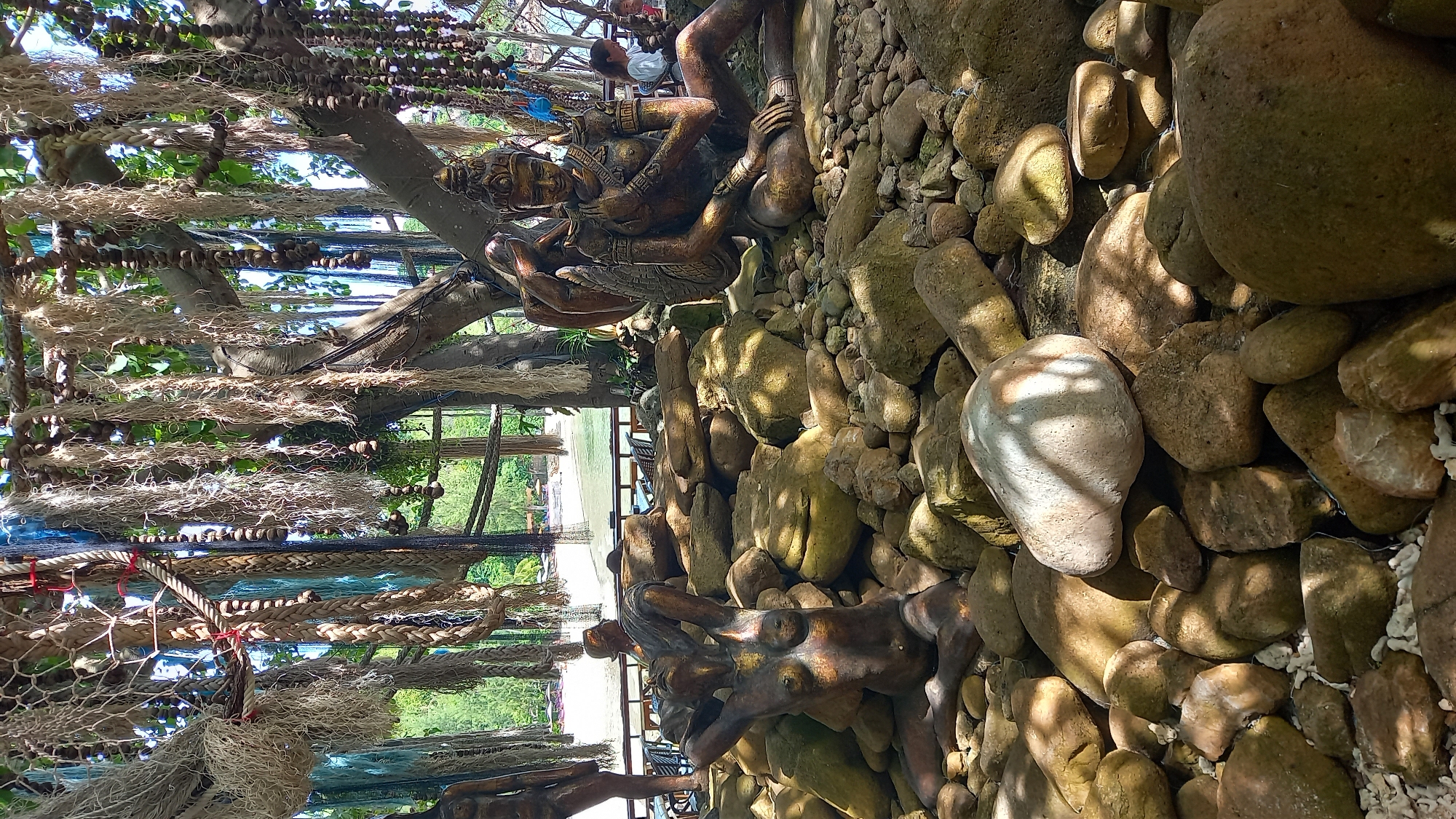 The image captures a serene outdoor scene in Koh Phangan, Thailand, featuring several bronze statues amidst a natural landscape. In the foreground, a collection of weathered rocks forms the base for two prominent statues. One statue depicts a reclining nude female figure with her hands behind her head, partially obscured by rocks. To the right, another statue portrays a figure in a seated, meditative pose, adorned with traditional jewelry and a headdress.
Overhead, a large tree with exposed roots supports a canopy of natural decorations, including fishing nets, ropes, and hanging strings of beads or seed pods, suggesting an artistic or decorative arrangement. In the background, a body of water, likely the sea, is visible, with a sandy beach and distant treeline. Some tables and chairs are set up near the water, indicating a recreational or dining area. A few individuals can be seen in the background, one woman is seated at a table, and another person is partially visible. The overall atmosphere is peaceful and artistic, blending natural elements with human-made sculptures.