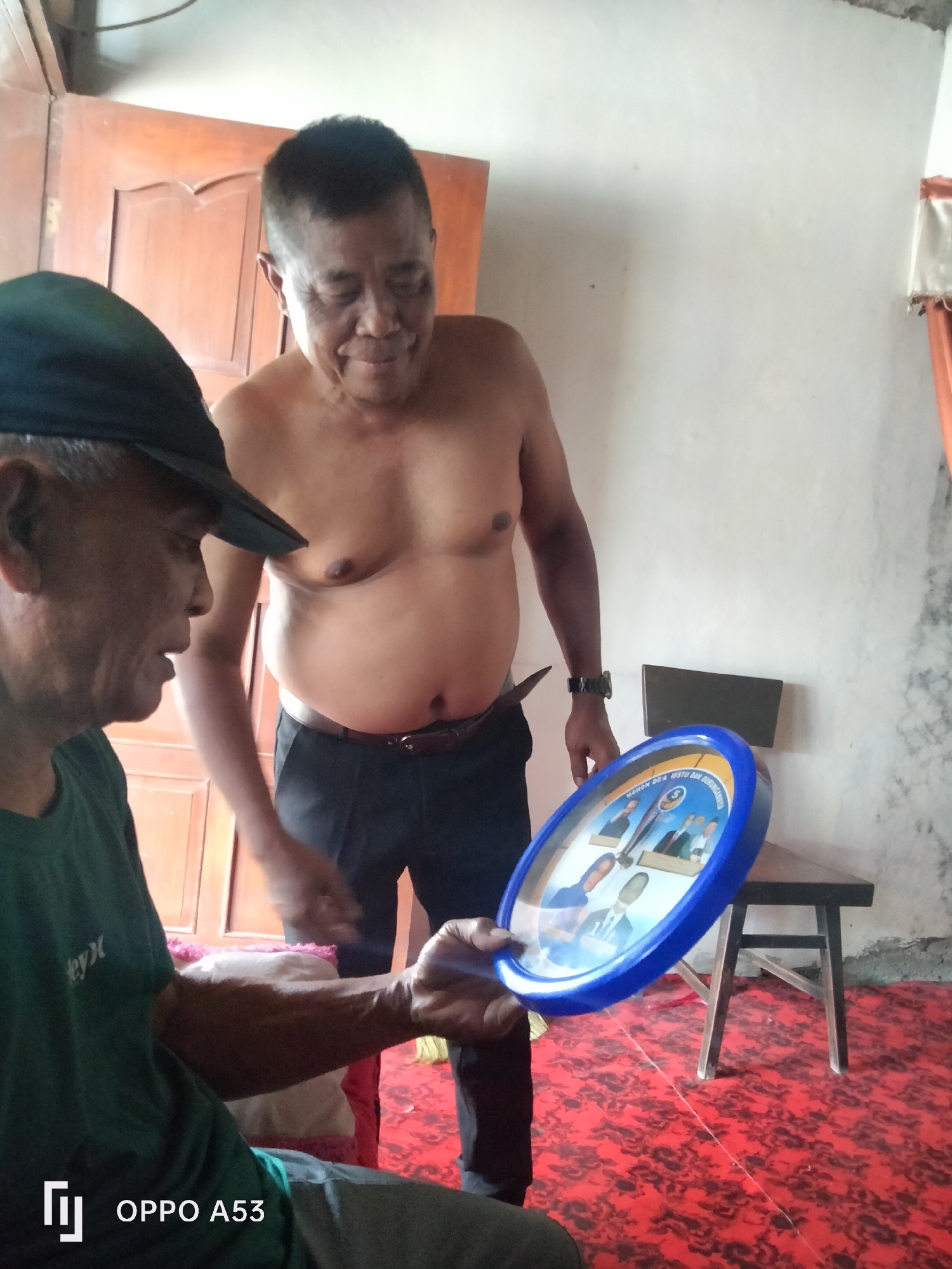 Two adult males are indoors, possibly in the city of Karangan, Indonesia. The male in the foreground, wearing a dark green t-shirt and a dark baseball cap, is holding a blue-rimmed circular clock in his left hand. The clock face displays four individuals in formal attire, two men and two women, along with some text. This male's right arm is extended, holding the clock. He is looking downwards and to his right.
The second male, shirtless and wearing black pants with a dark belt, stands to the right and slightly behind the first man. He is looking downwards towards the clock and appears to be smiling. He has a watch on his left wrist.
The environment includes a light-colored wall, a wooden door in the background, and a red carpet with a black floral pattern covering the floor. A dark wooden stool or chair is visible in the mid-ground to the right. The lighting suggests an indoor setting with natural light. The camera is positioned low, looking up slightly at the two men. The image has a watermark in the bottom left corner that reads "OPPO A53".