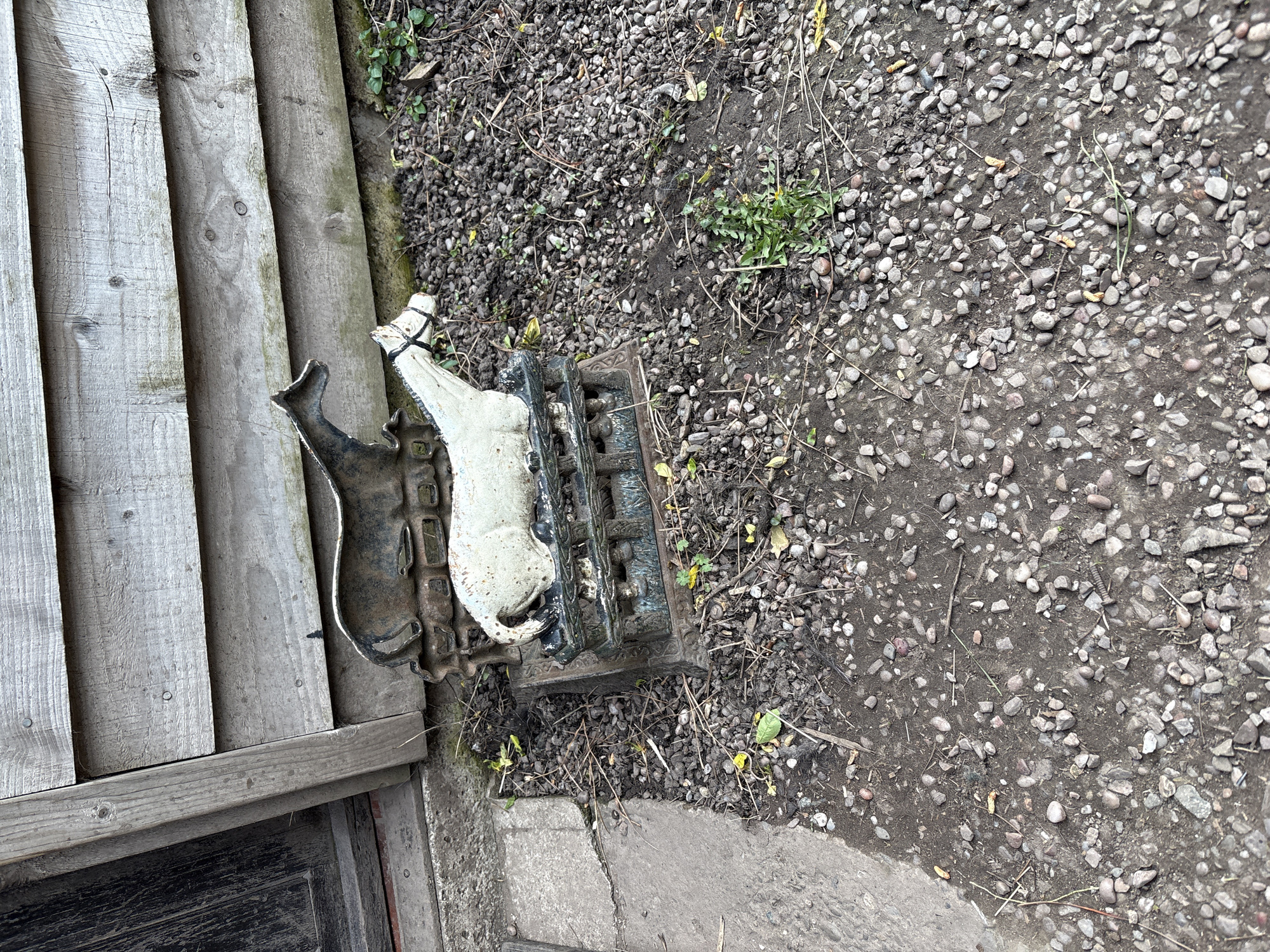 The image shows a cast iron boot scraper with a white horse figure attached, lying next to a weathered wooden wall. The surrounding ground is covered with small stones and gravel, suggesting an outdoor environment, possibly a garden or yard. The location is assumed to be Pulverbatch, United Kingdom. No people or actions are depicted.