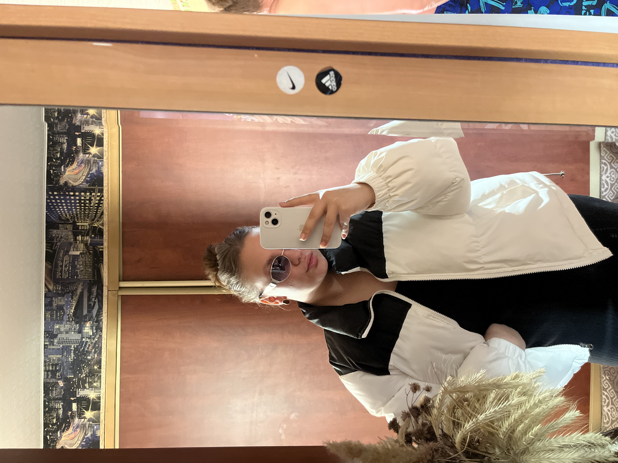 A person wearing a white and black jacket is taking a selfie in front of a mirror. The mirror's frame has a picture of a city on the left, possibly a cityscape. There are two stickers on the top of the mirror frame: a Nike swoosh and an Adidas logo. The person has sunglasses on and is holding a smartphone. Part of a dried flower arrangement is visible at the bottom of the image. The background seen through the mirror includes a plain wall. The location is presumed to be in Ольховка, Russia, based on context.