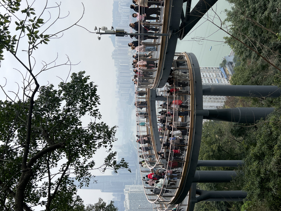 An elevated, curved walkway crowded with people traverses a green, tree-filled landscape. Supporting columns anchor the structure, with a backdrop revealing a misty cityscape and a river or lake. Bare branches frame the left, and dense foliage flanks the right side. Security cameras are visible atop the walkway.