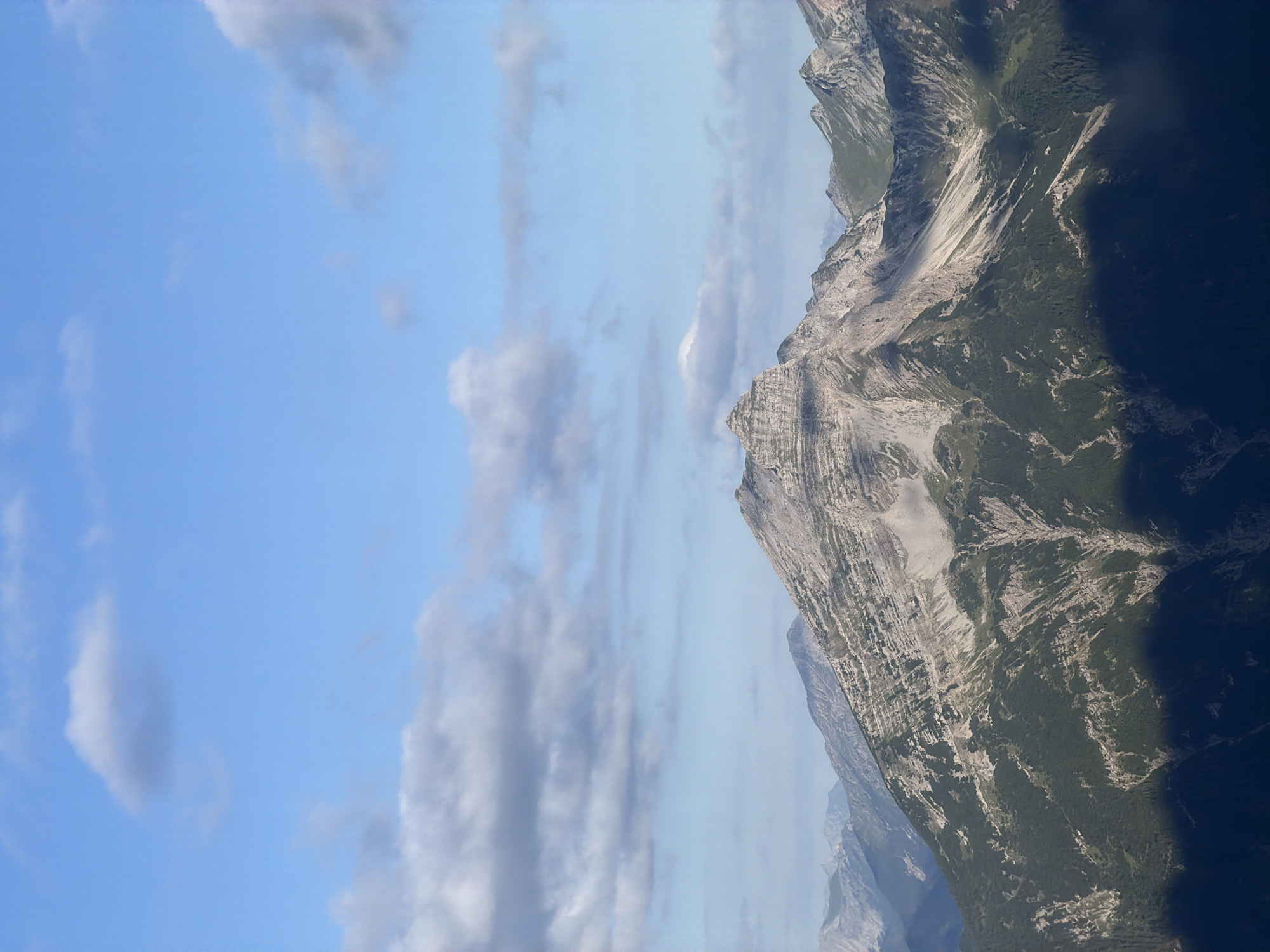 The image shows a panoramic view of a mountain range under a partly cloudy sky. The foreground is dominated by dark shadows cast by the mountains. In the mid-ground, a large, rugged mountain with visible rock strata and patches of green vegetation rises prominently. It features steep slopes, gullies, and scree. Further into the distance, a series of less distinct mountain peaks extend, with some appearing green and others more rocky. The sky is a vibrant blue with scattered white and gray clouds, some of which are elongated and wispy. The lighting suggests it is daytime, possibly late afternoon, given the long shadows. There are no people or man-made objects visible in this natural landscape. The scene depicts a remote and untamed mountain environment.