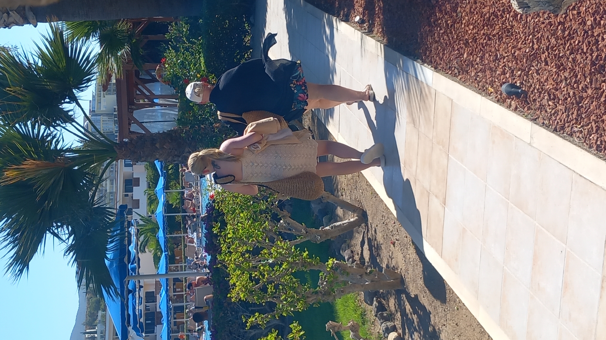 Two women are walking on a paved pathway at a resort in Kos, Greece. The woman in the foreground on the left is wearing a light-colored, knee-length dress and carrying a woven bag. The woman on the right is wearing a dark top and a floral-patterned skirt, also carrying a woven bag. They are walking away from the camera, towards the right side of the image. In the background, there are lounge chairs, blue umbrellas, and a swimming pool. Palm trees and lush green foliage are visible, characteristic of a Mediterranean climate. The pathway is bordered by a bed of red mulch. The sky is clear and blue.