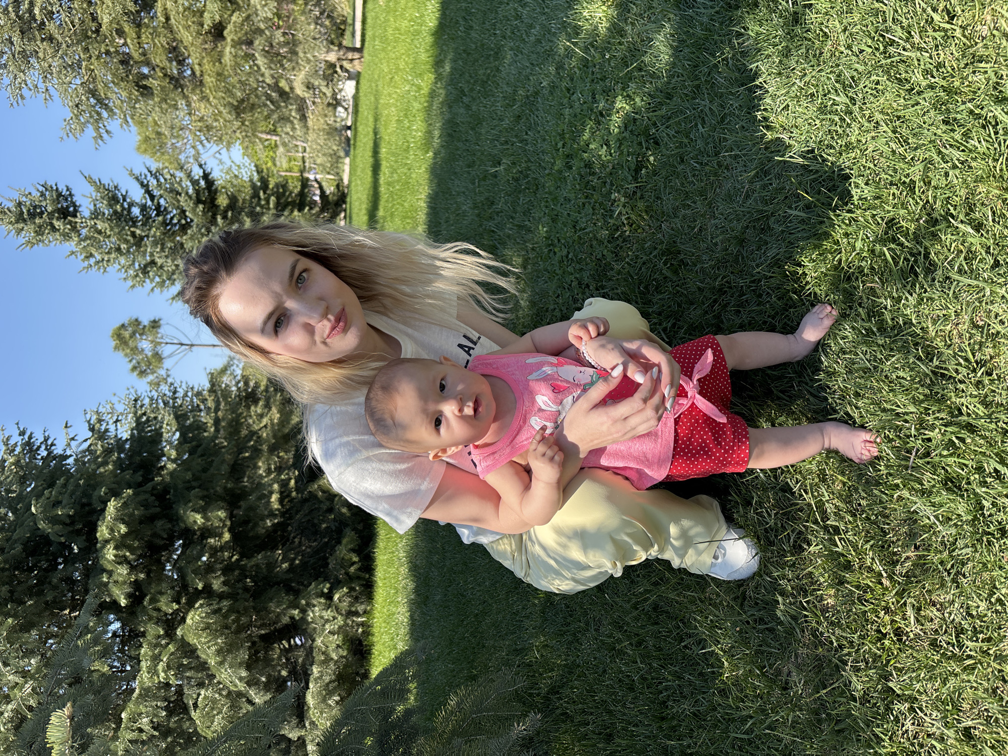 A fair-skinned woman with blonde hair is seated cross-legged on a lawn, holding a baby. The baby is wearing a pink top and red polka-dot bottoms with a pink bow. The woman is wearing a white t-shirt and pale yellow pants, with white sneakers. They are positioned on bright green grass with dappled sunlight. In the background are trees and a blue sky. The image was taken in Krasnodar, Russia.