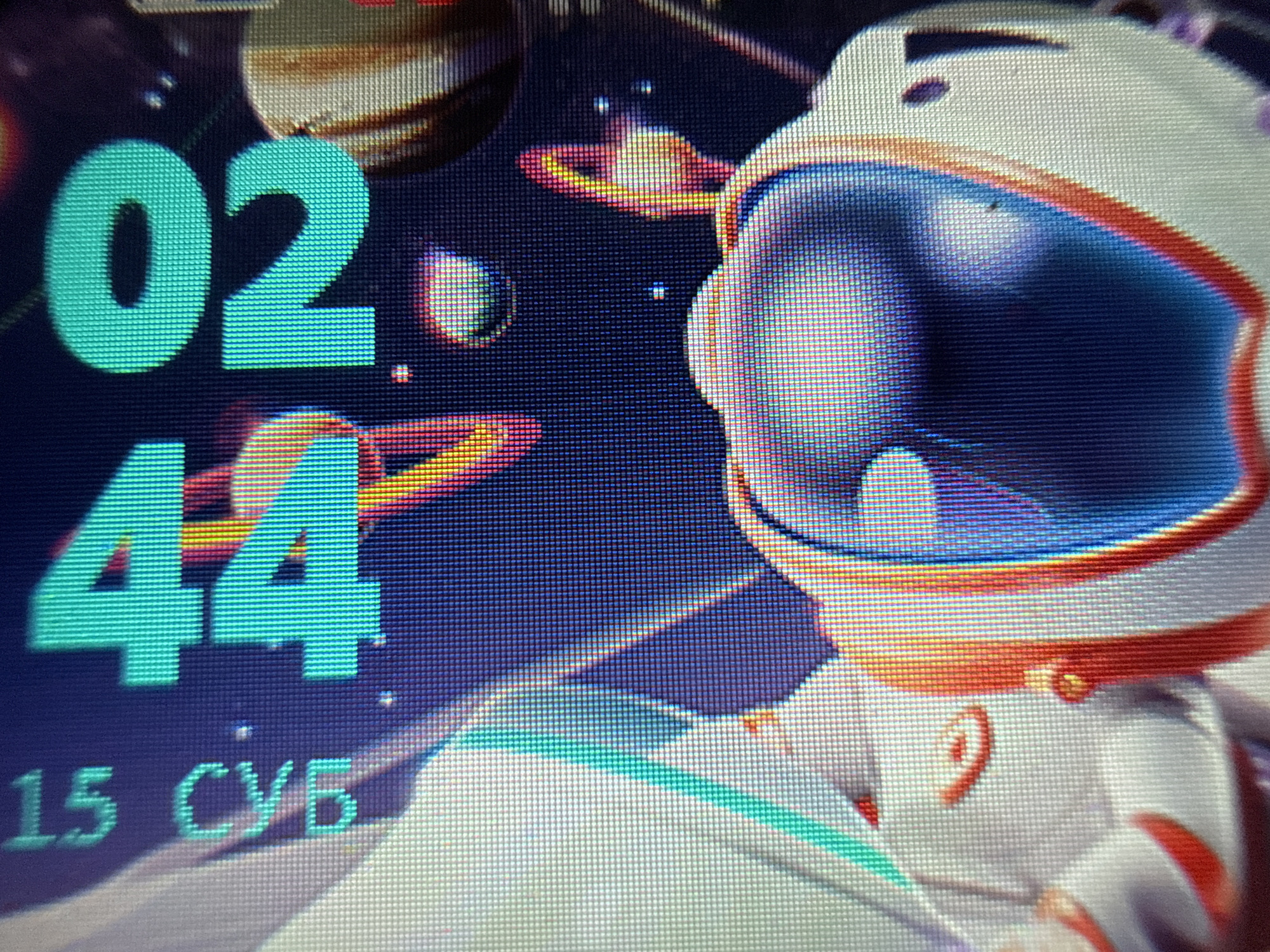 The image shows a screen displaying a digital clock set to 02:44 and the date "15 СУБ". An astronaut illustration is also visible on the screen, alongside depictions of planets and celestial bodies. There is no information available to suggest this is in the city of Kosh, Armenia.